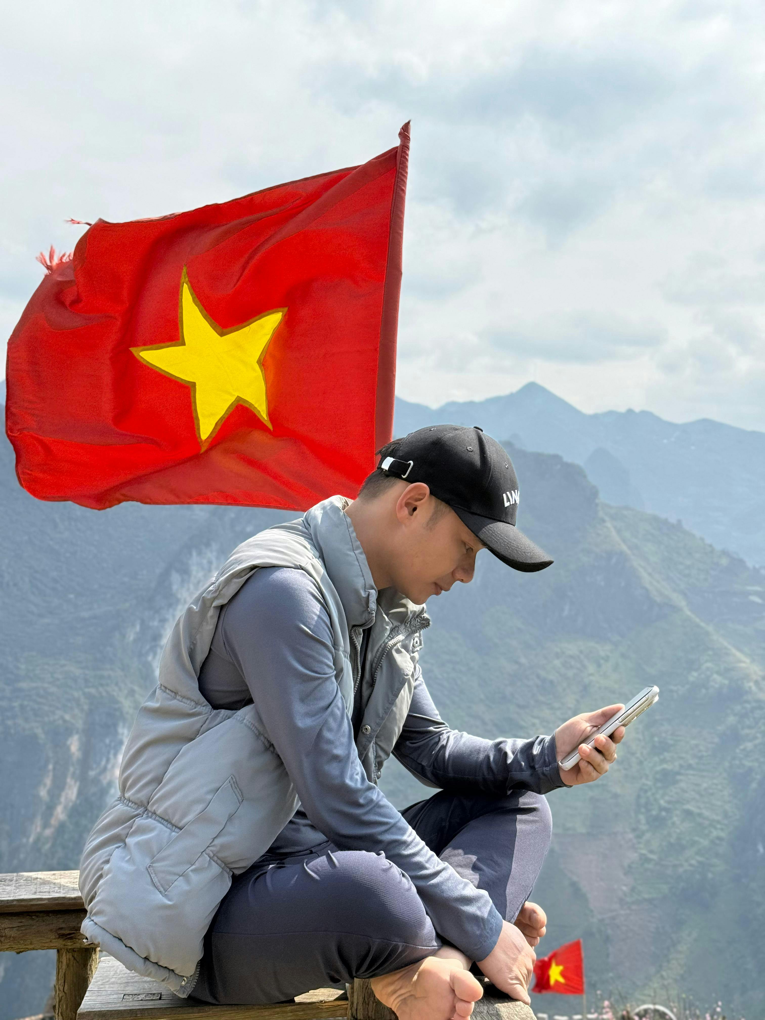 Gratuit Un homme est assis à l'extérieur, utilisant un smartphone, avec un drapeau vietnamien aux couleurs vives en arrière-plan. Photos