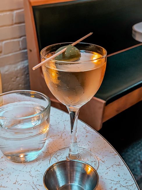 A chilled martini glass filled with a cloudy, pale gold dirty martini, three brine-soaked olives on a pick resting across the rim.