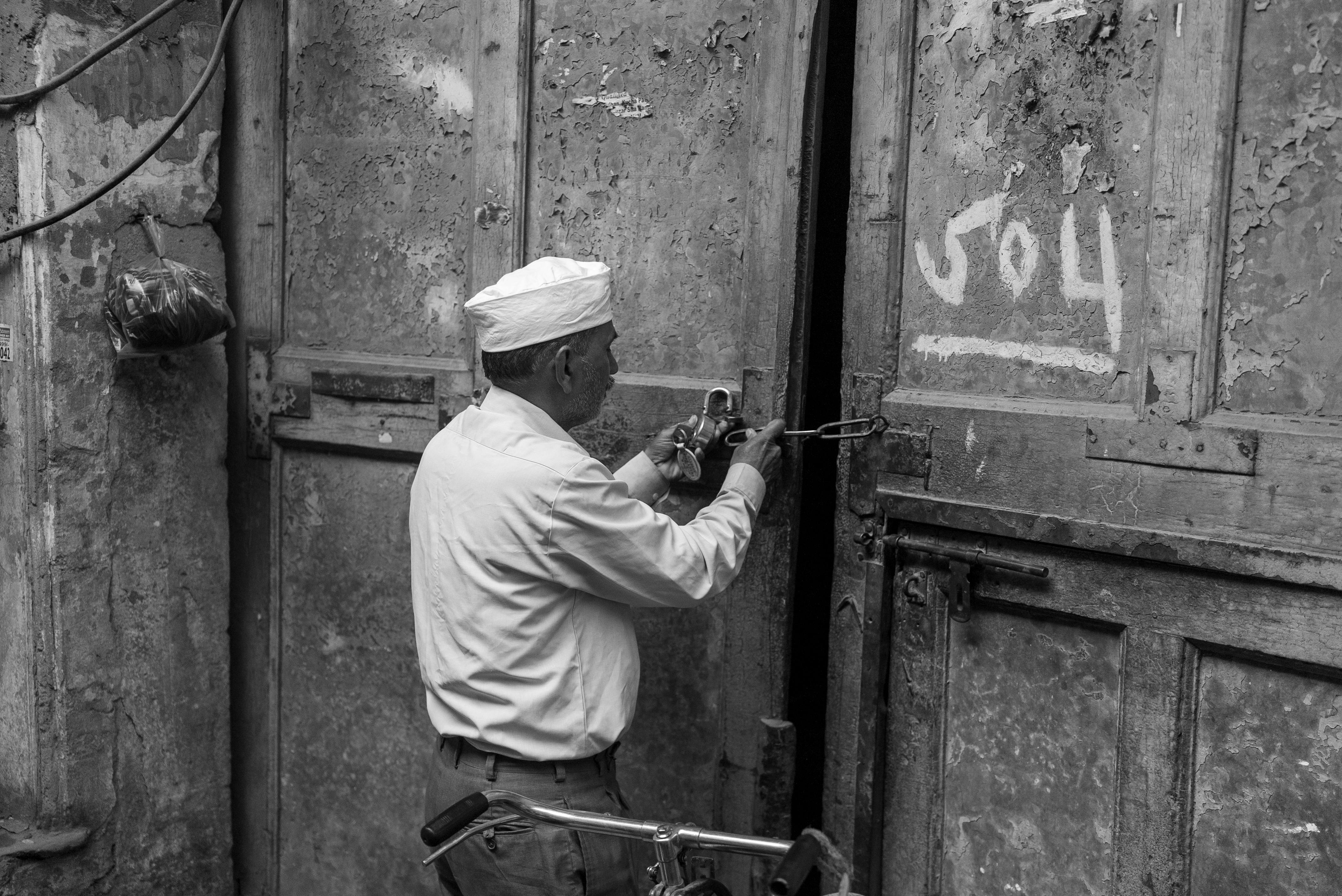 Miễn phí Một người đàn ông lớn tuổi trong trang phục truyền thống đang khóa một cánh cửa gỗ cũ kỹ trong một khu phố đô thị bụi bặm. Ảnh lưu trữ