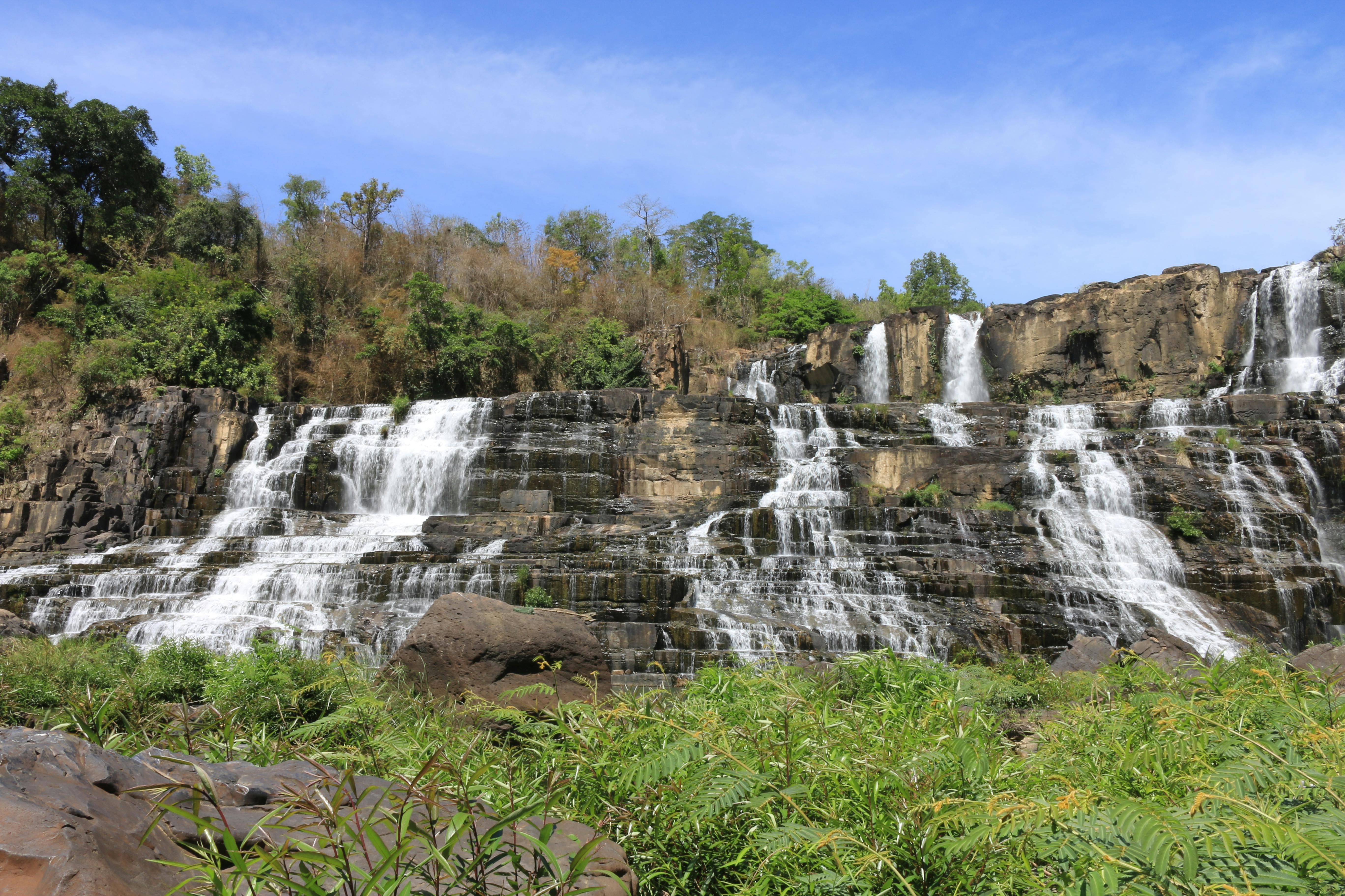 Free A stunning waterfall cascades over rocks surrounded by vibrant green foliage under a clear blue sky. Stock Photo
