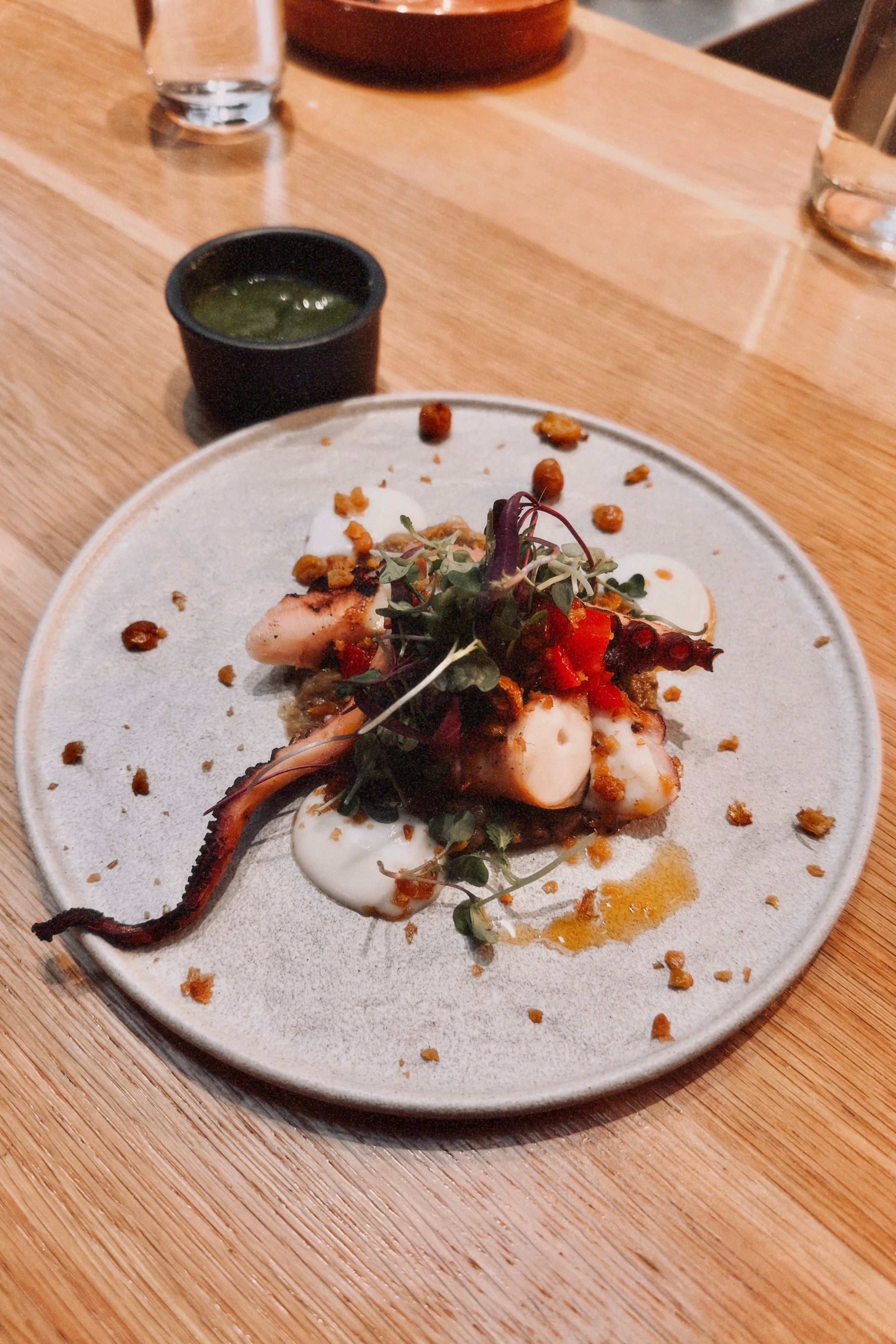 A beautifully plated gourmet octopus dish with vibrant garnishes and green sauce on a wooden table.