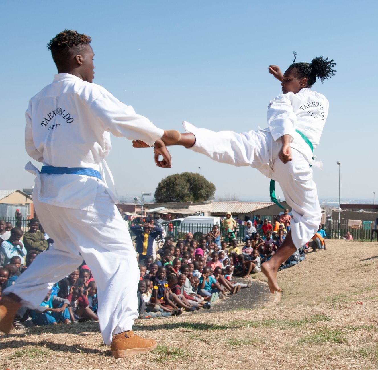 Two martial artists performing a dynamic Taekwondo demonstration outdoors, engaging an audience.