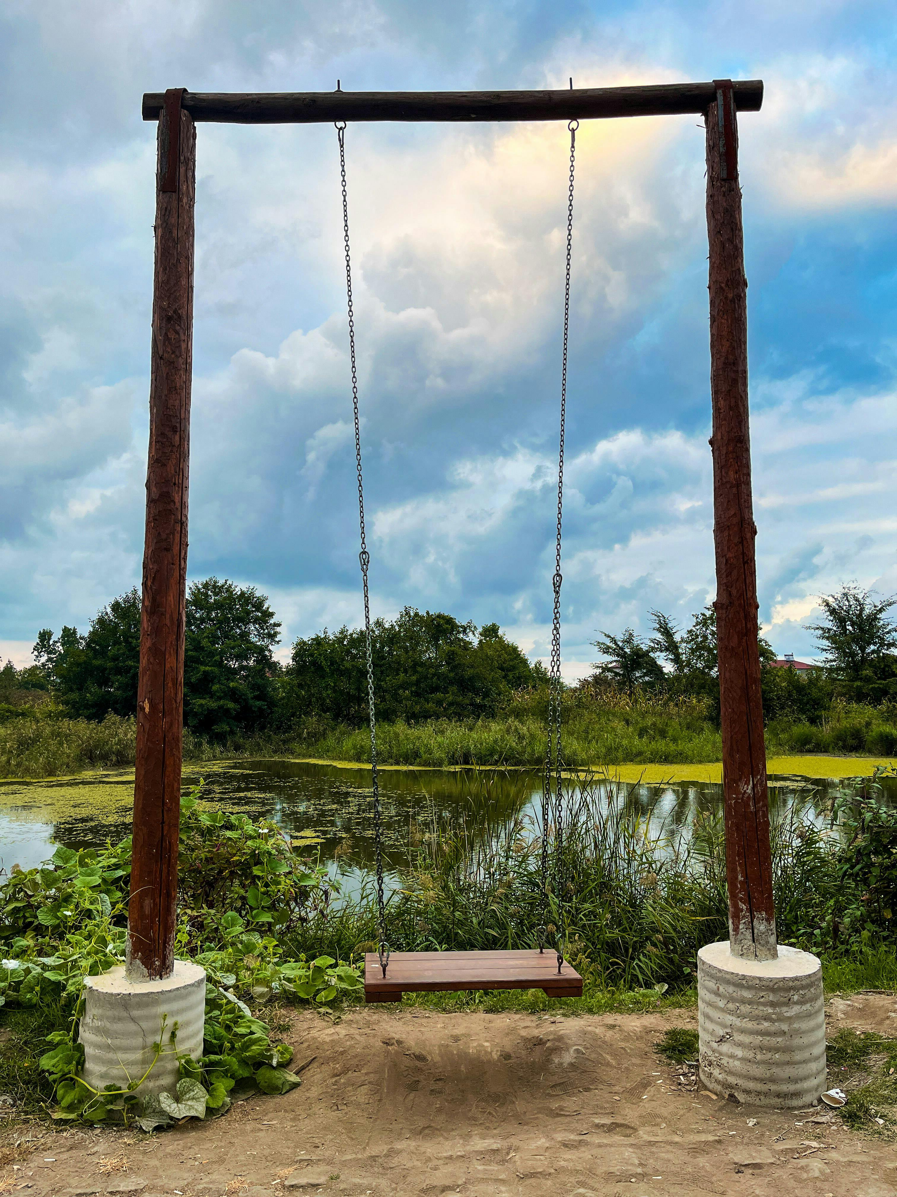 Kostenlos Eine Holzschaukel vor der Kulisse eines idyllischen Teichs, umgeben von üppigem Grün. Stock-Foto