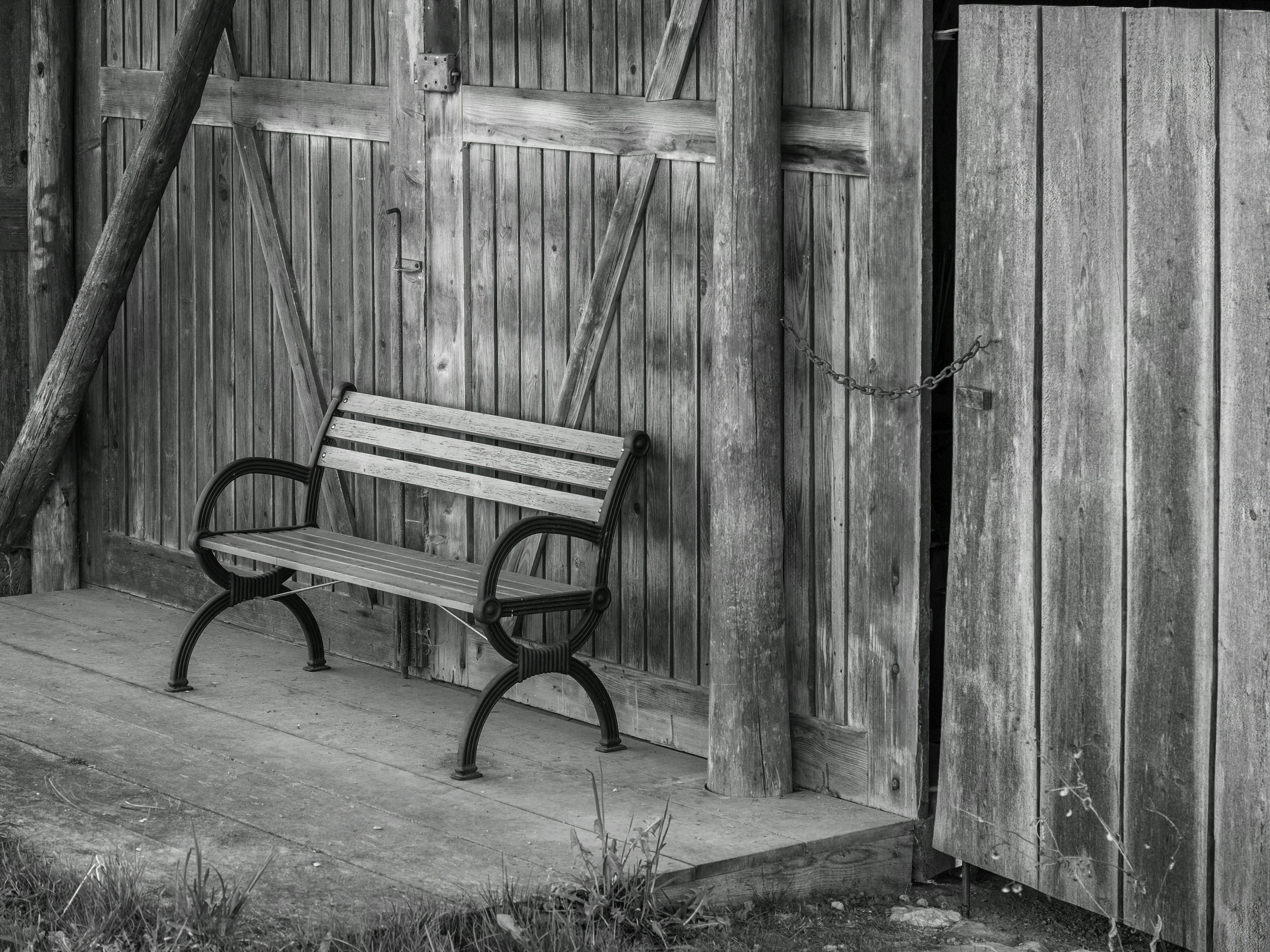 bezplatná Černobílý obrázek historické dřevěné lavičky před omšelou stodolou. Rustikální klid. Základová fotografie