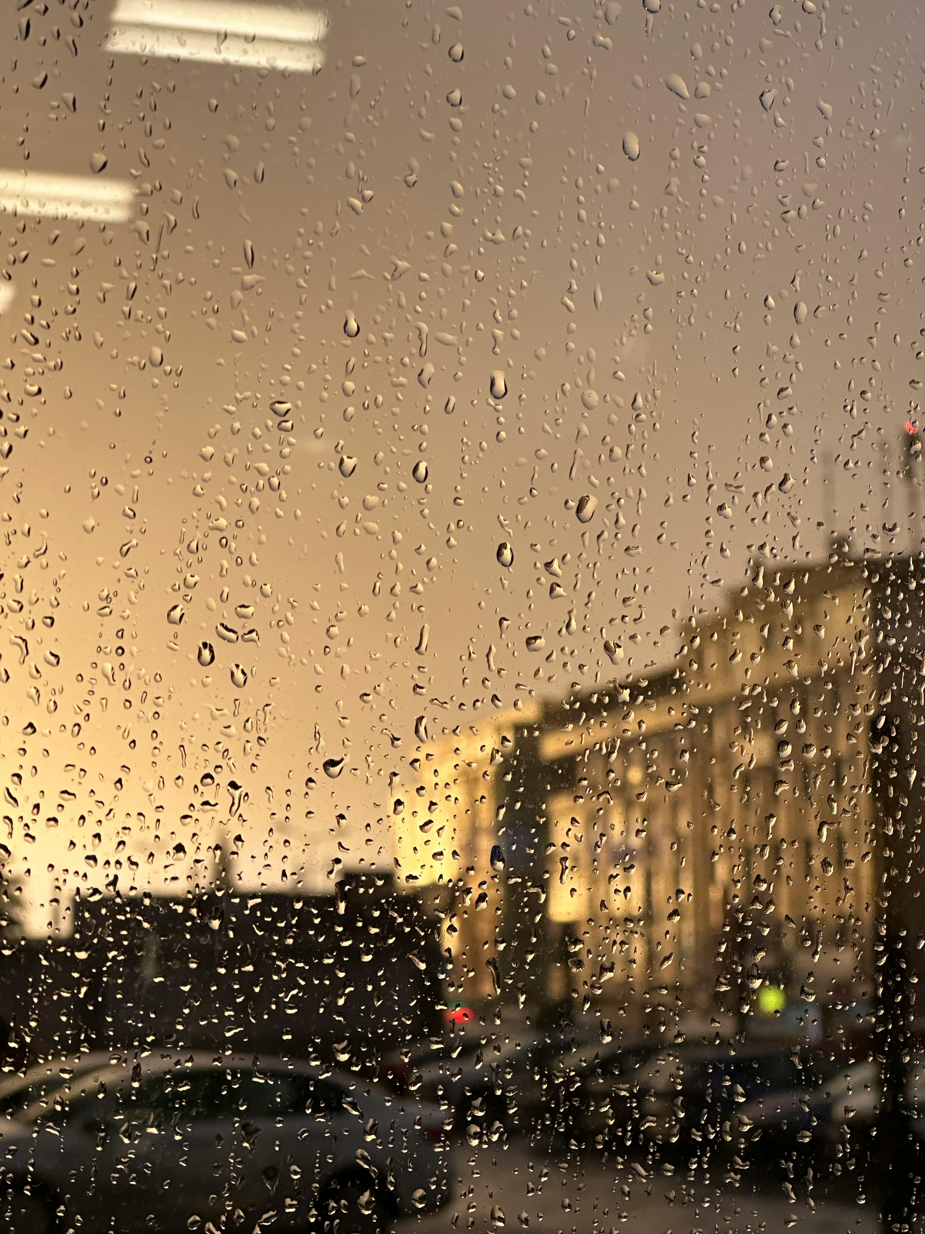 Kostenlos Regentropfen auf einem Fenster vor einer verschwommenen Stadtlandschaft im Hintergrund, aufgenommen in der Abenddämmerung, zeigen eine dramatische, stimmungsvolle Szene. Stock-Foto