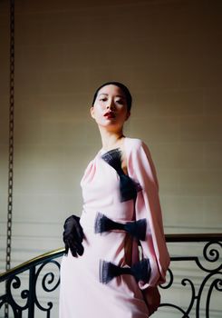 Stylish woman poses in pink dress with black accents and gloves by ornate staircase.