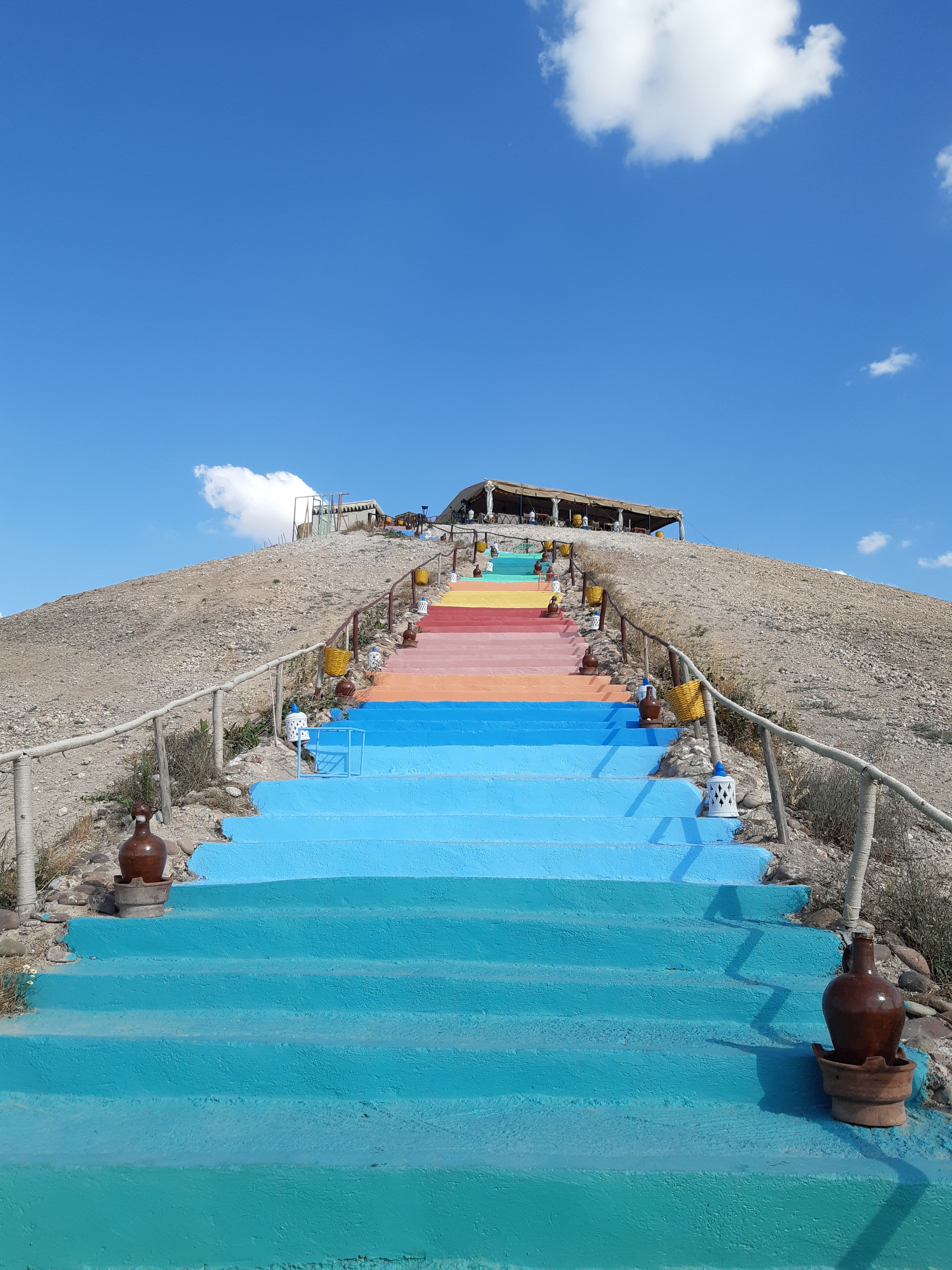 Vibrant rainbow stairs lead up a hill in the Moroccan desert under a bright blue sky.