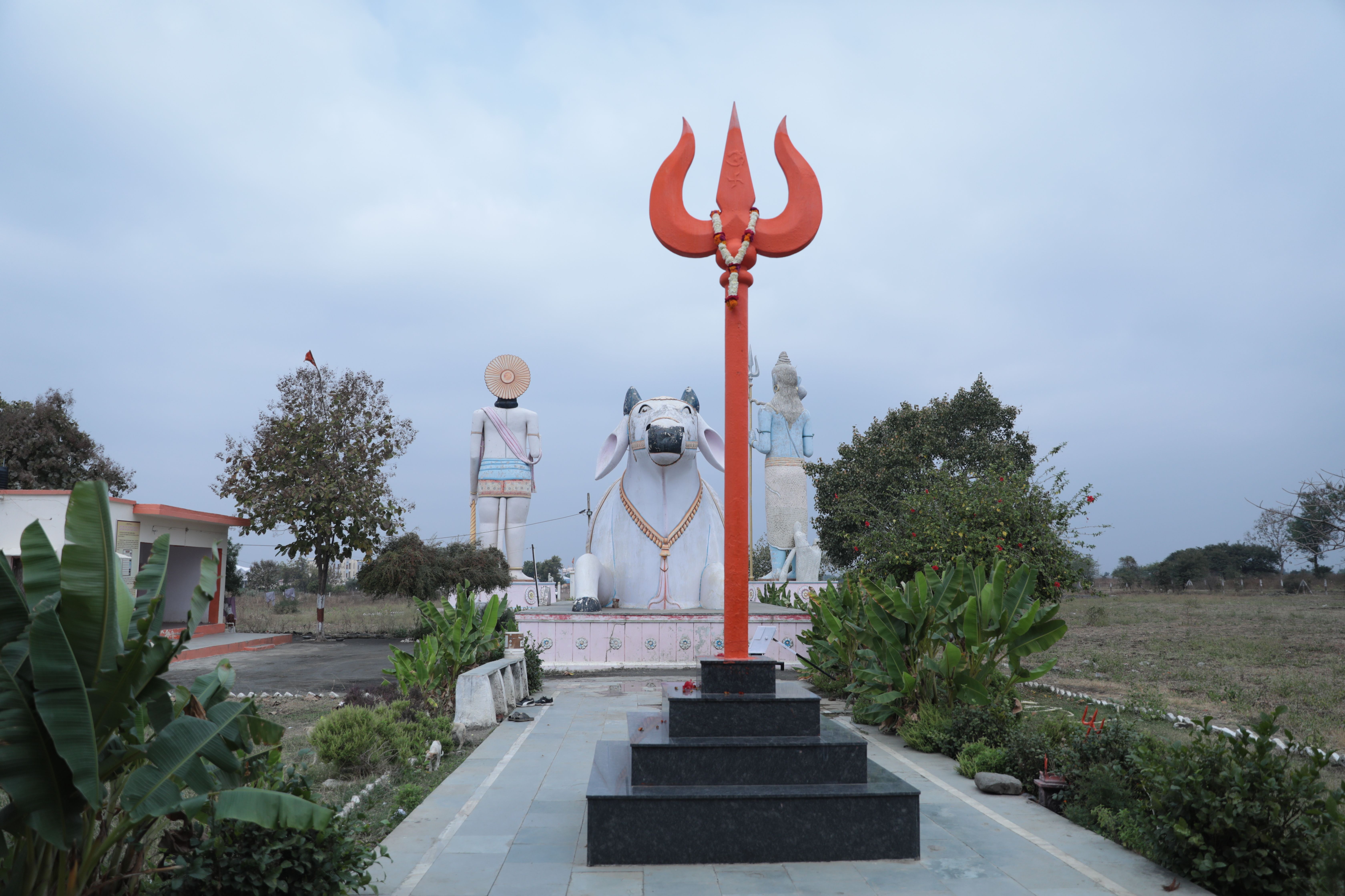 Peaceful Hindu temple setting featuring a large Nandi statue and Trishul against a cloudy sky.
