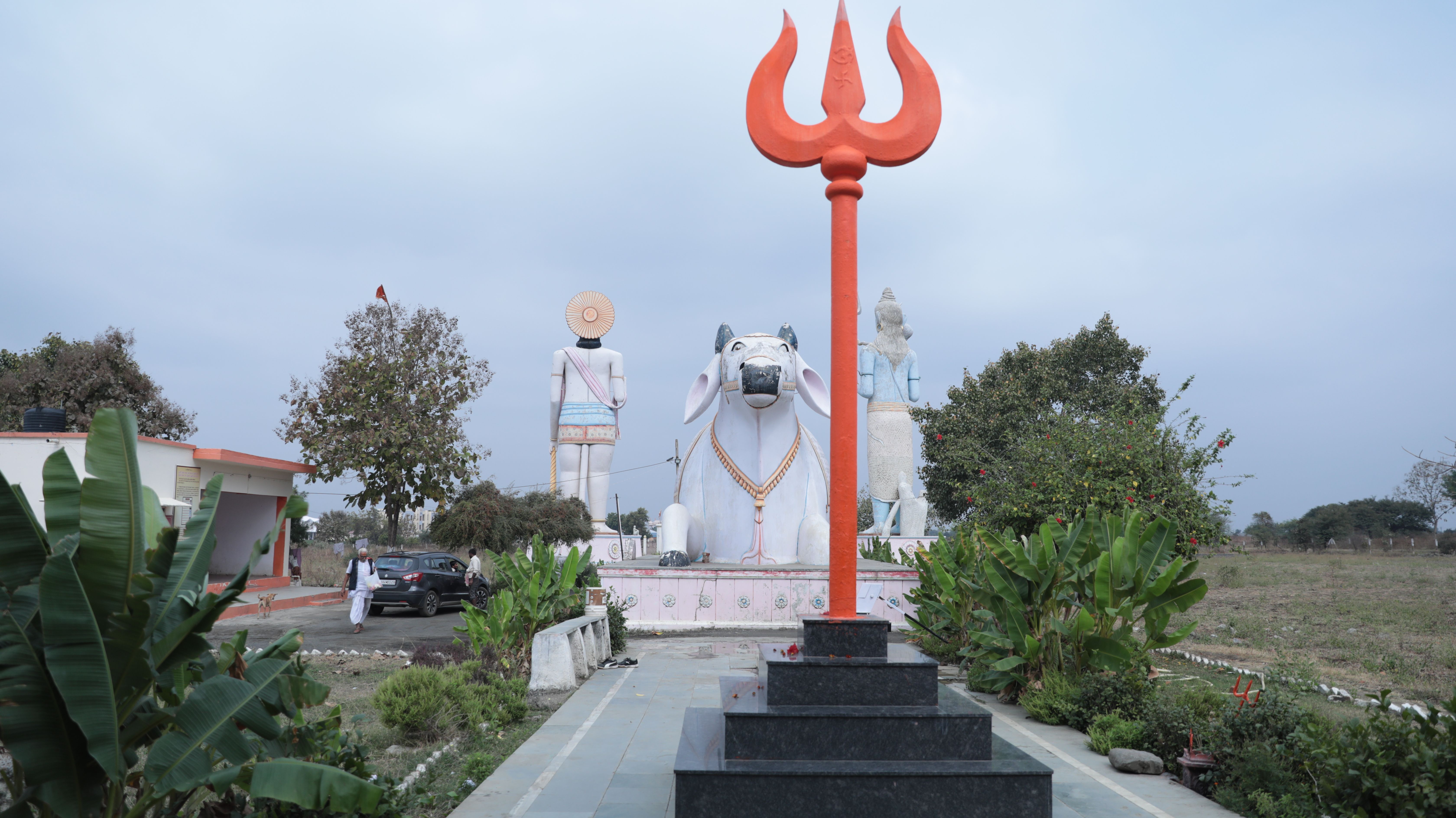 A serene temple courtyard featuring a towering trident and Nandi statue, surrounded by lush greenery.