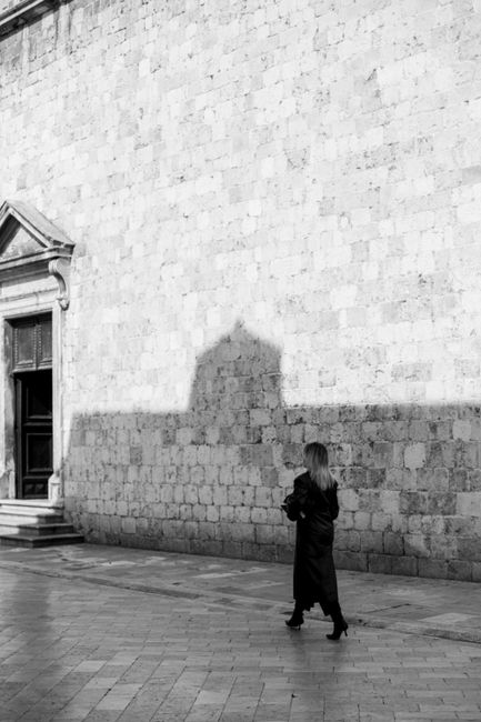 Black and white photo of a woman in silhouette walking against an ancient stone wall, casting a dramatic shadow.
