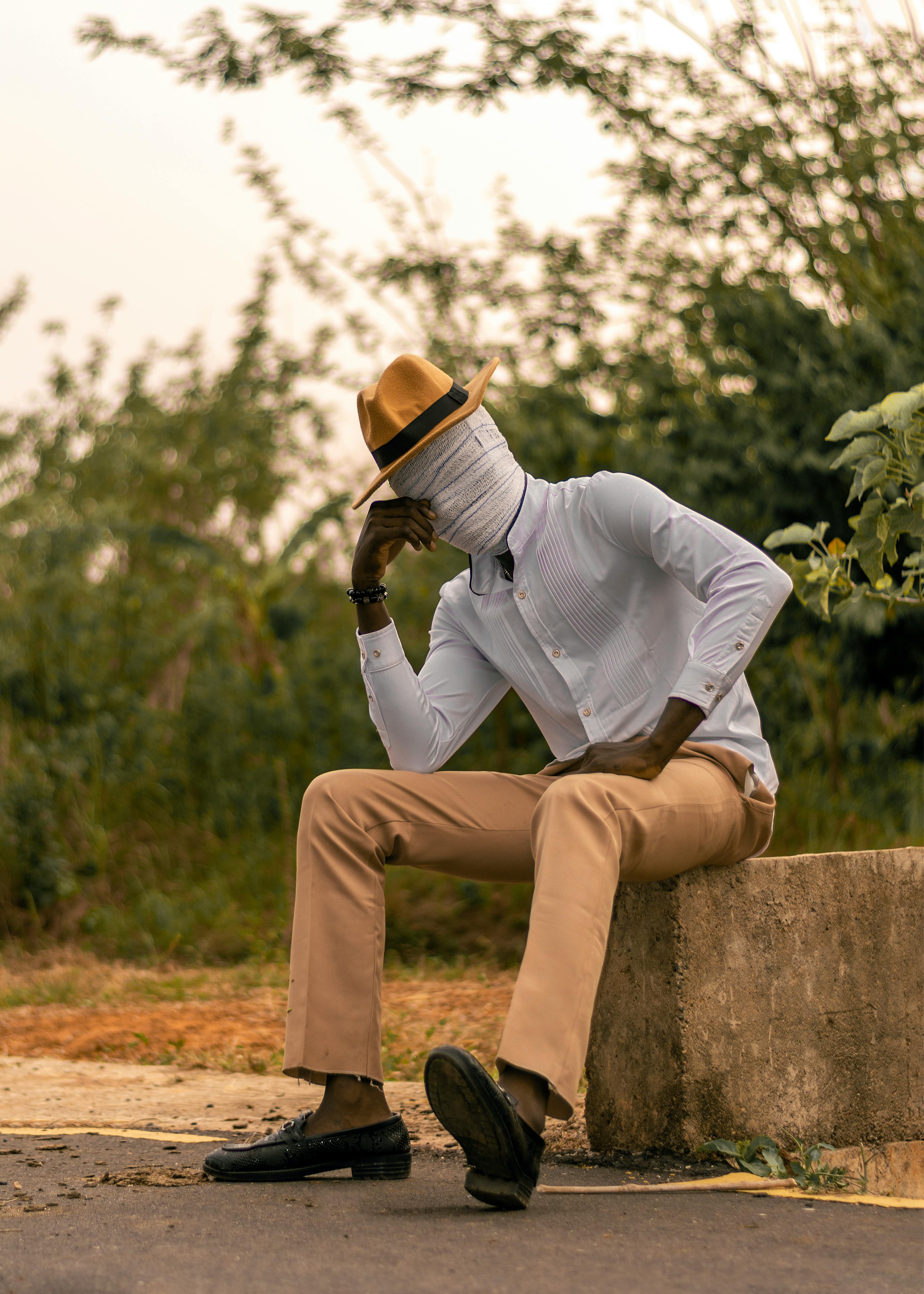 免費 一名身著時尚服裝、臉部被遮蓋的男子坐在自然環境中的路邊。 圖庫相片