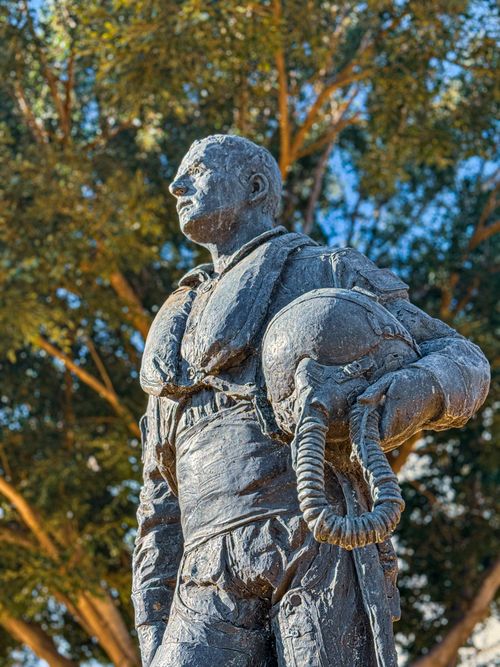 Free A bronze statue depicting a pilot with a helmet under a tree, in a sunny outdoor setting. Stock Photo