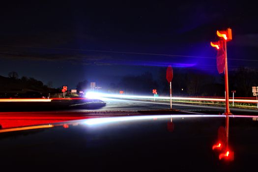 Long exposure of vehicle light trails at a rural intersection with stop signs and colorful reflections.