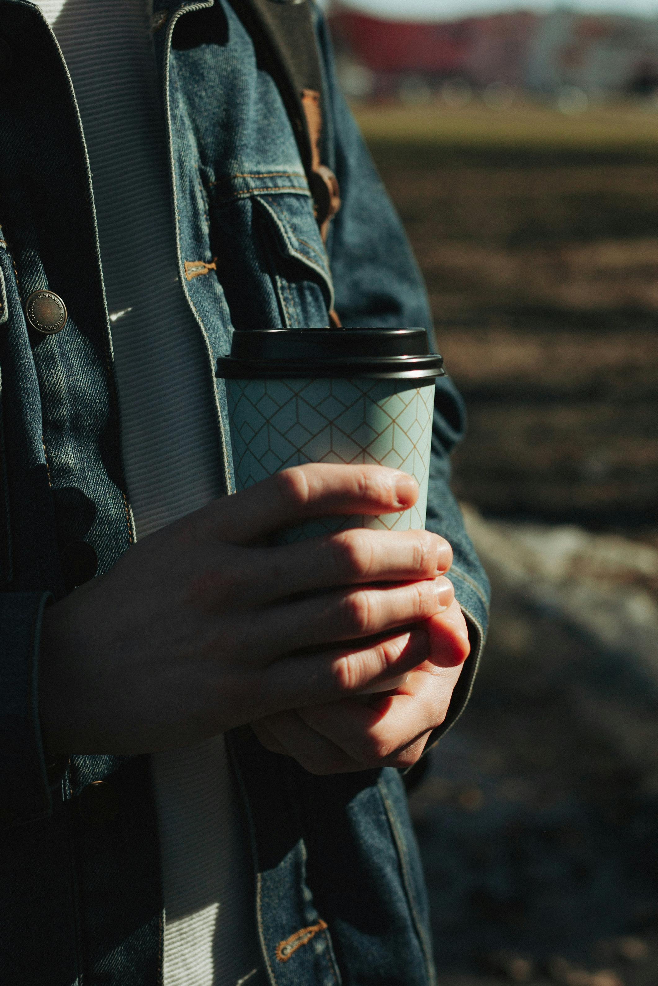 bezplatná Osoba držící šálek kávy venku za slunečného dne, ležérně oblečená v džínách. Základová fotografie