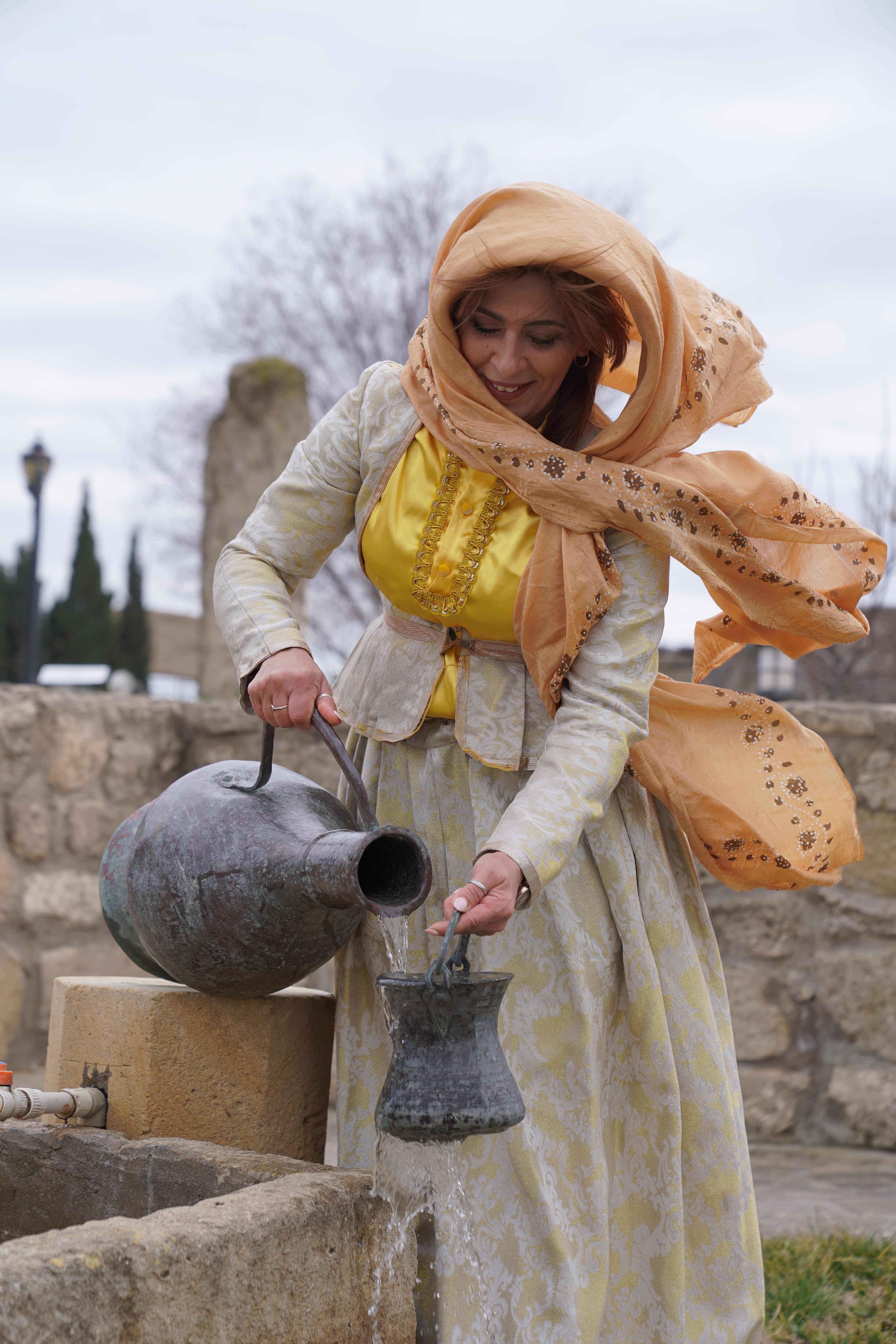 A woman in traditional attire pouring water from a metal jug outdoors, embodying cultural heritage.