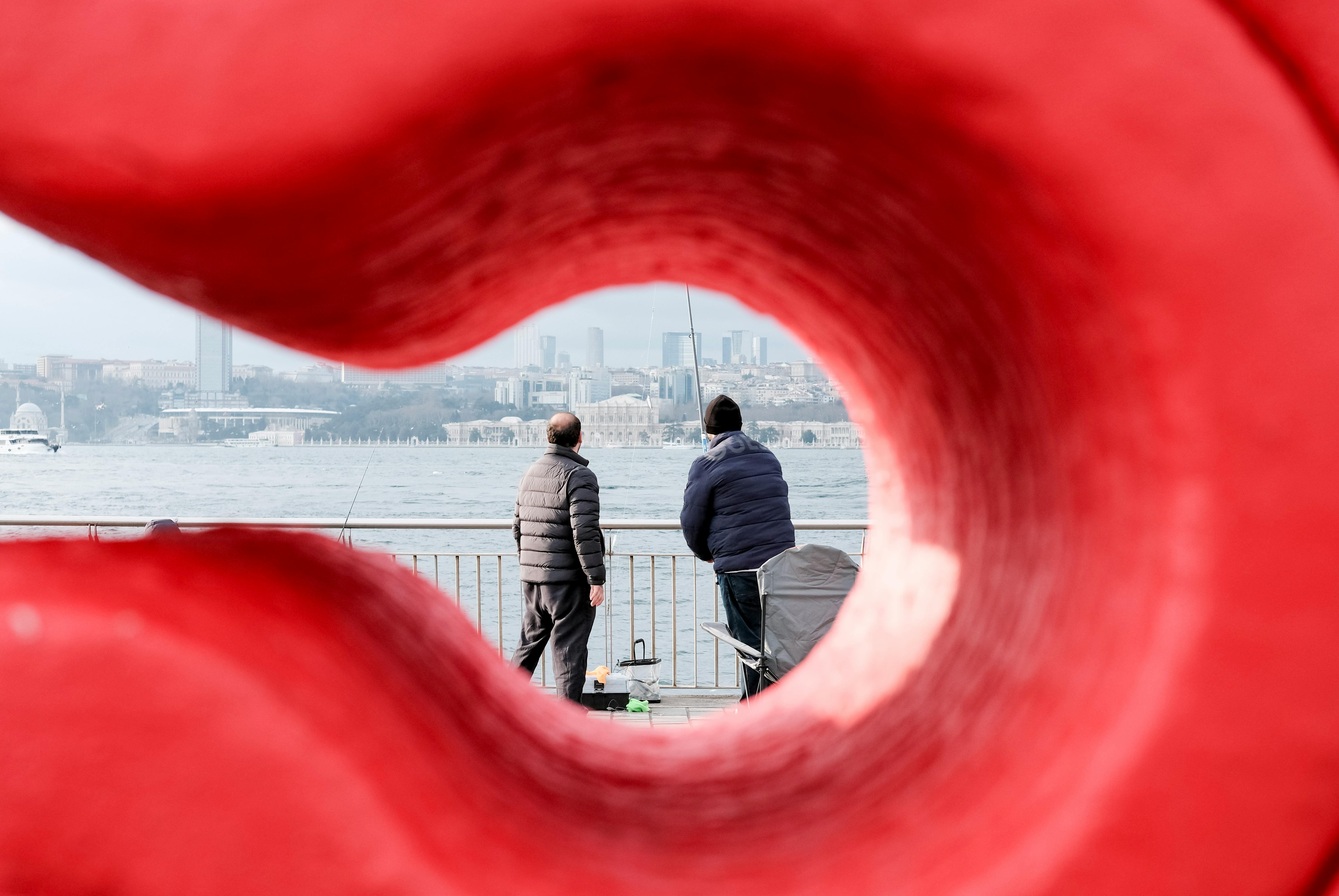 Kostnadsfria Ett unikt perspektiv på två män som fiskar vid Bosporen, inramat av en röd skulptur i Istanbul. Stock foto