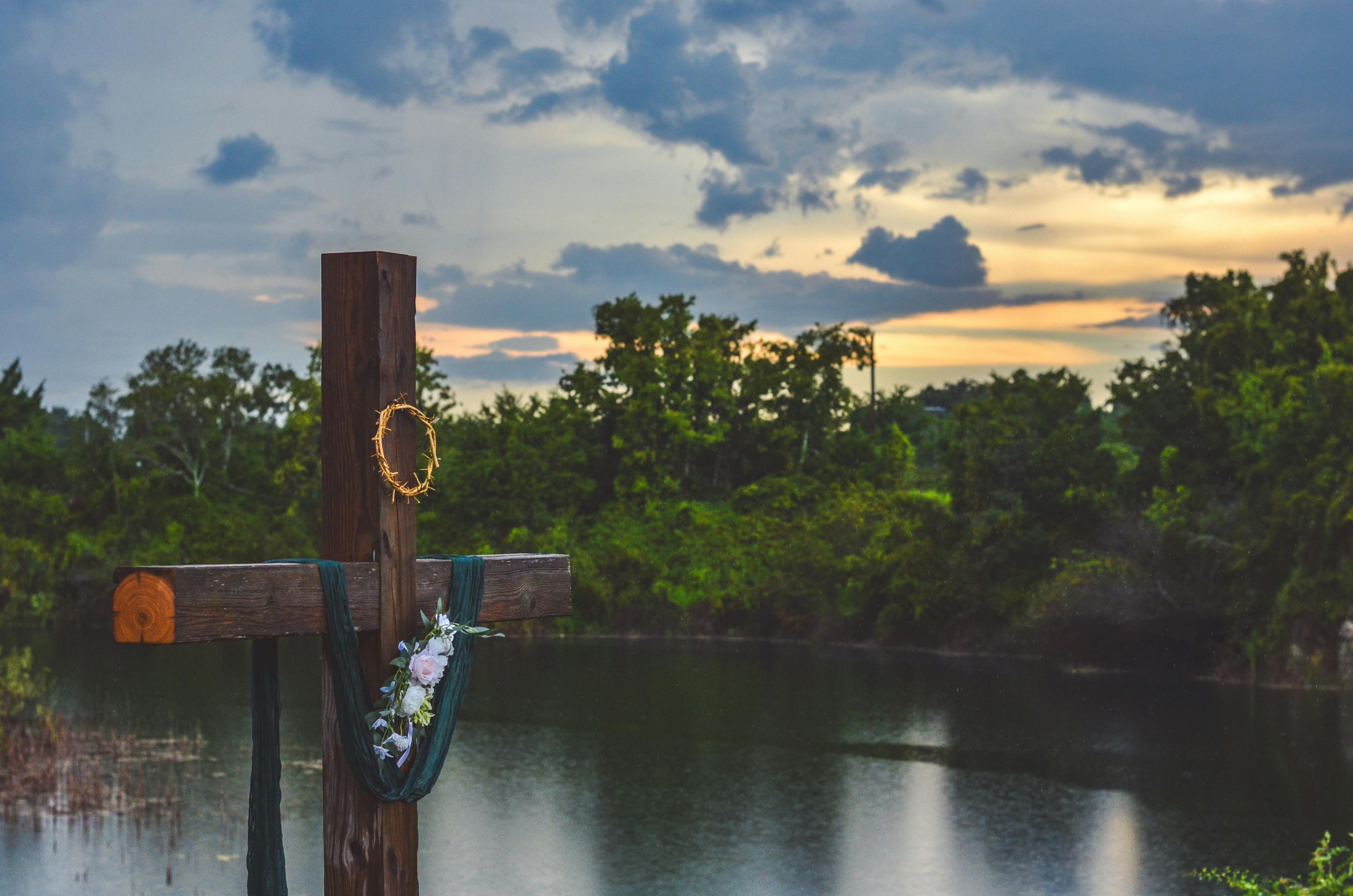 grátis Vista panorâmica de uma cruz de madeira adornada com uma coroa de espinhos e flores junto a um lago tranquilo ao pôr do sol. Foto profissional