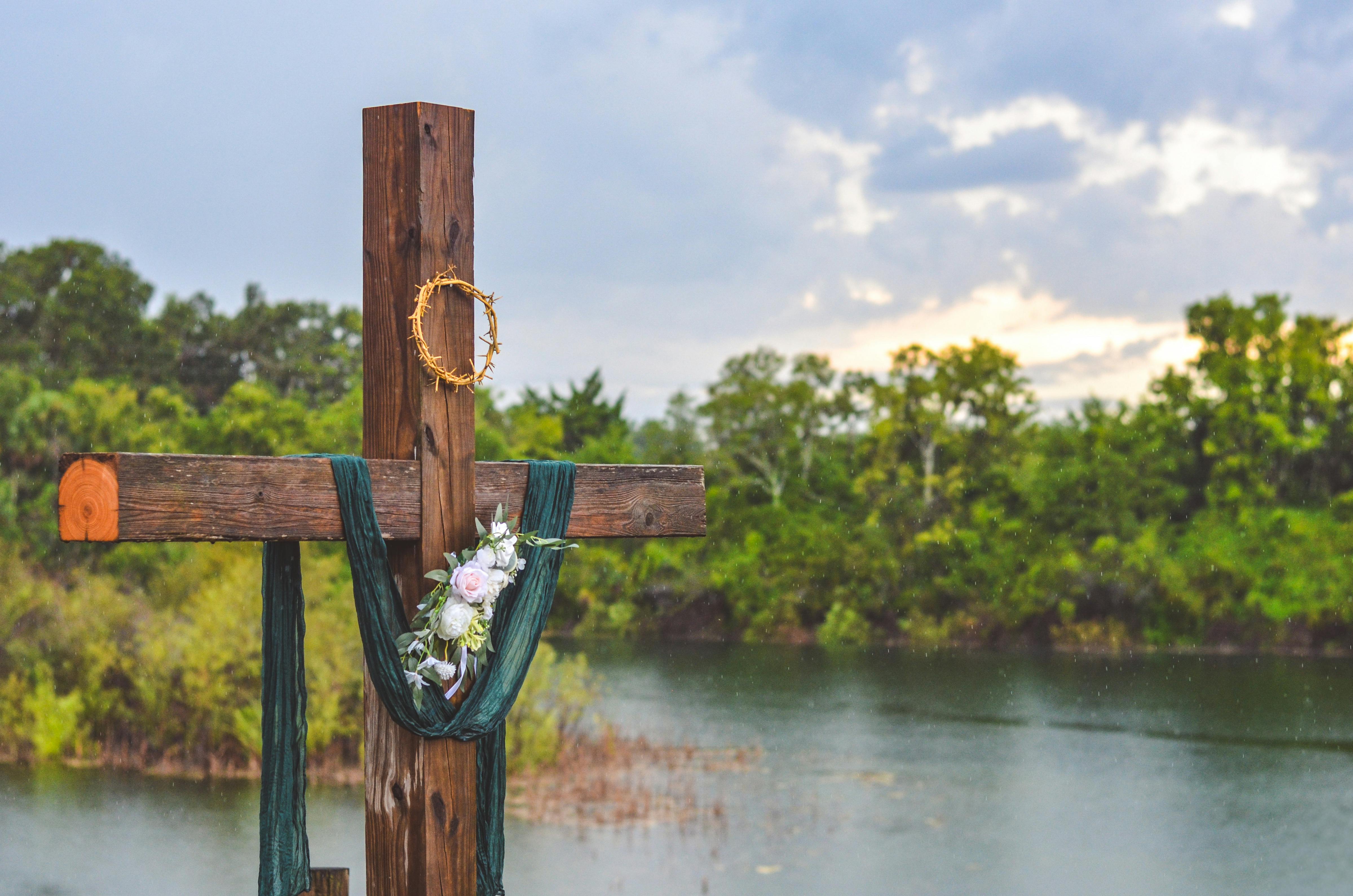 Δωρεάν Ένας ξύλινος σταυρός στολισμένος με λουλούδια και αγκάθινο στεφάνι δίπλα σε μια λίμνη κατά τη διάρκεια του ηλιοβασιλέματος, που συμβολίζει την ειρήνη και την πνευματικότητα. Φωτογραφία από στοκ φωτογραφιών