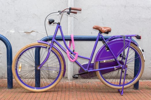 Vibrant purple bicycle securely locked to a blue rack against a brick path backdrop.