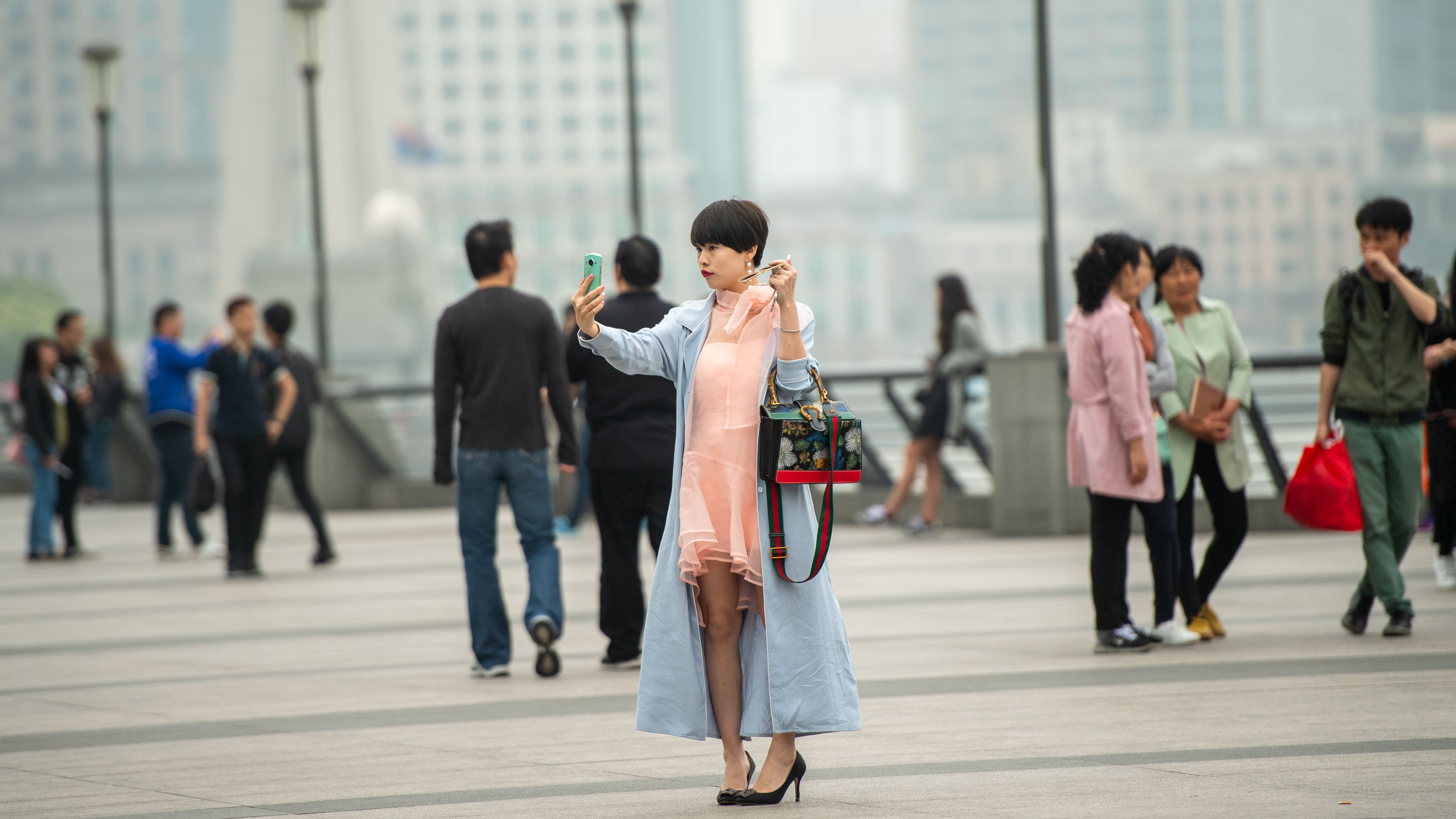 A fashionable woman captures a selfie on a busy Shanghai promenade, showing urban Chinese lifestyle