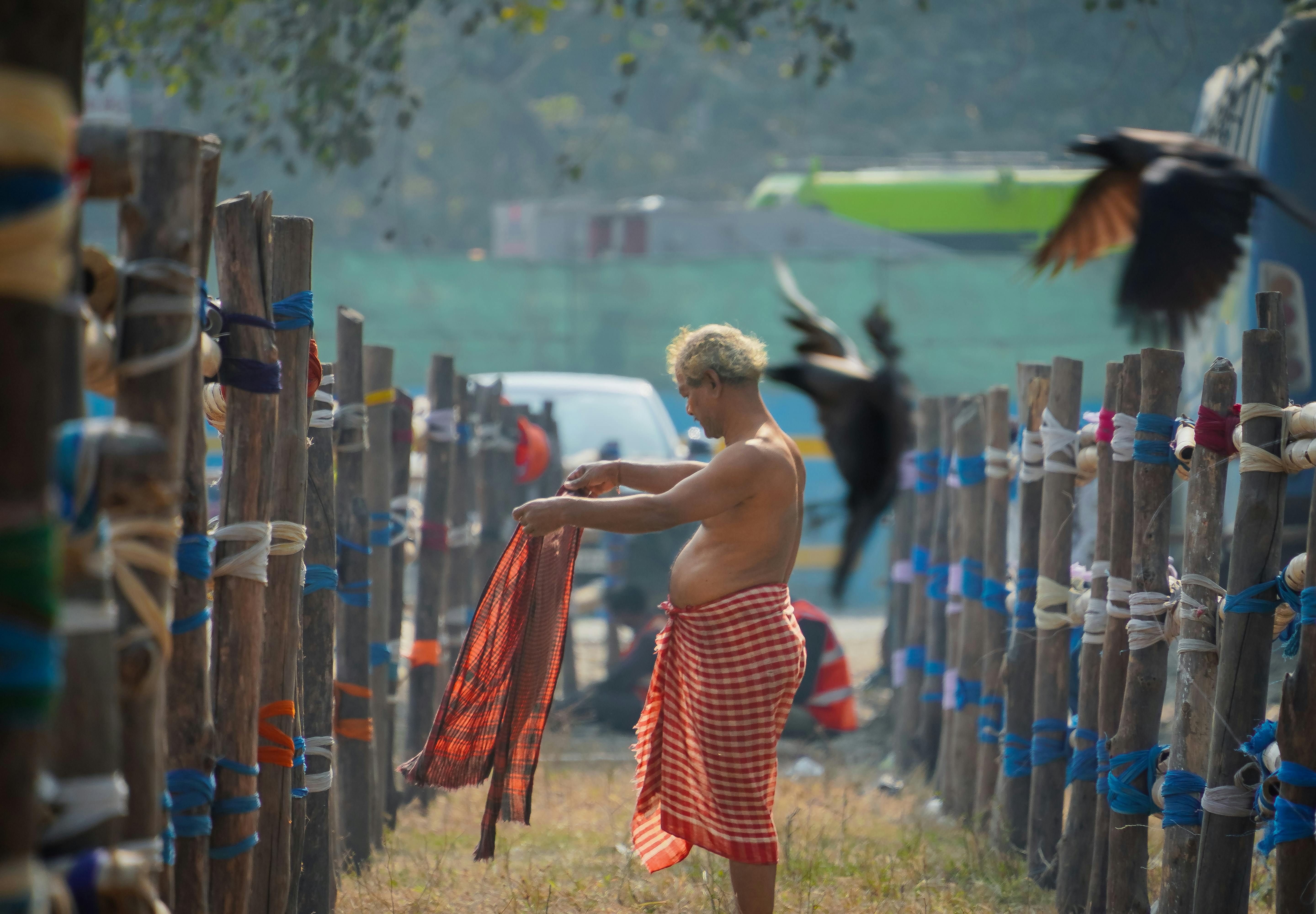 Gratuit Un bărbat împachetează o pânză tradițională afară, înconjurat de stâlpi de lemn. Păsările zboară în apropiere. Fotografie de stoc