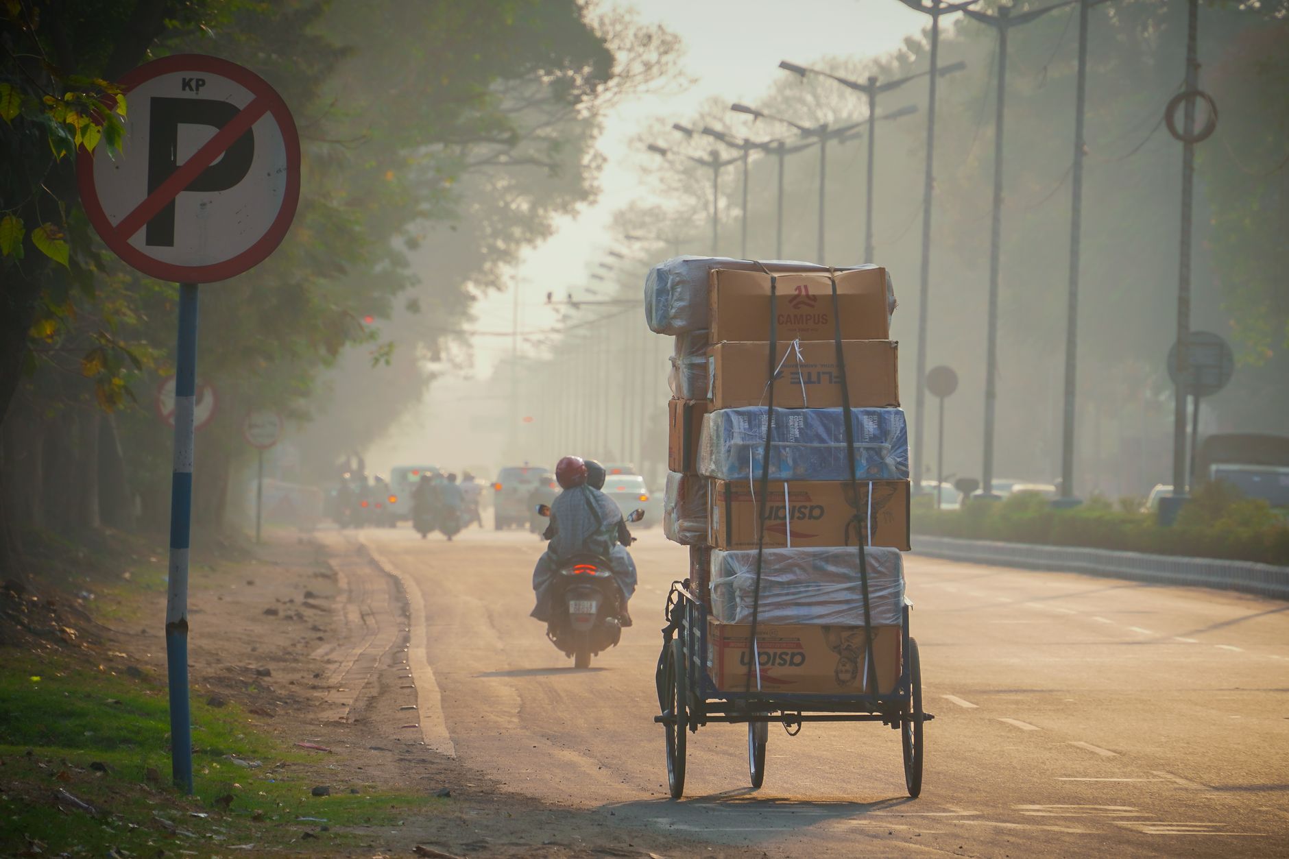 A delivery tricycle carrying packages on a misty Indian street during daylight with a no parking sign.