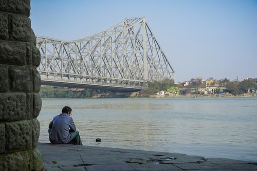 Free A man sits by the Hooghly River near the iconic Howrah Bridge in Kolkata, India, reflecting in solitude. Stock Photo