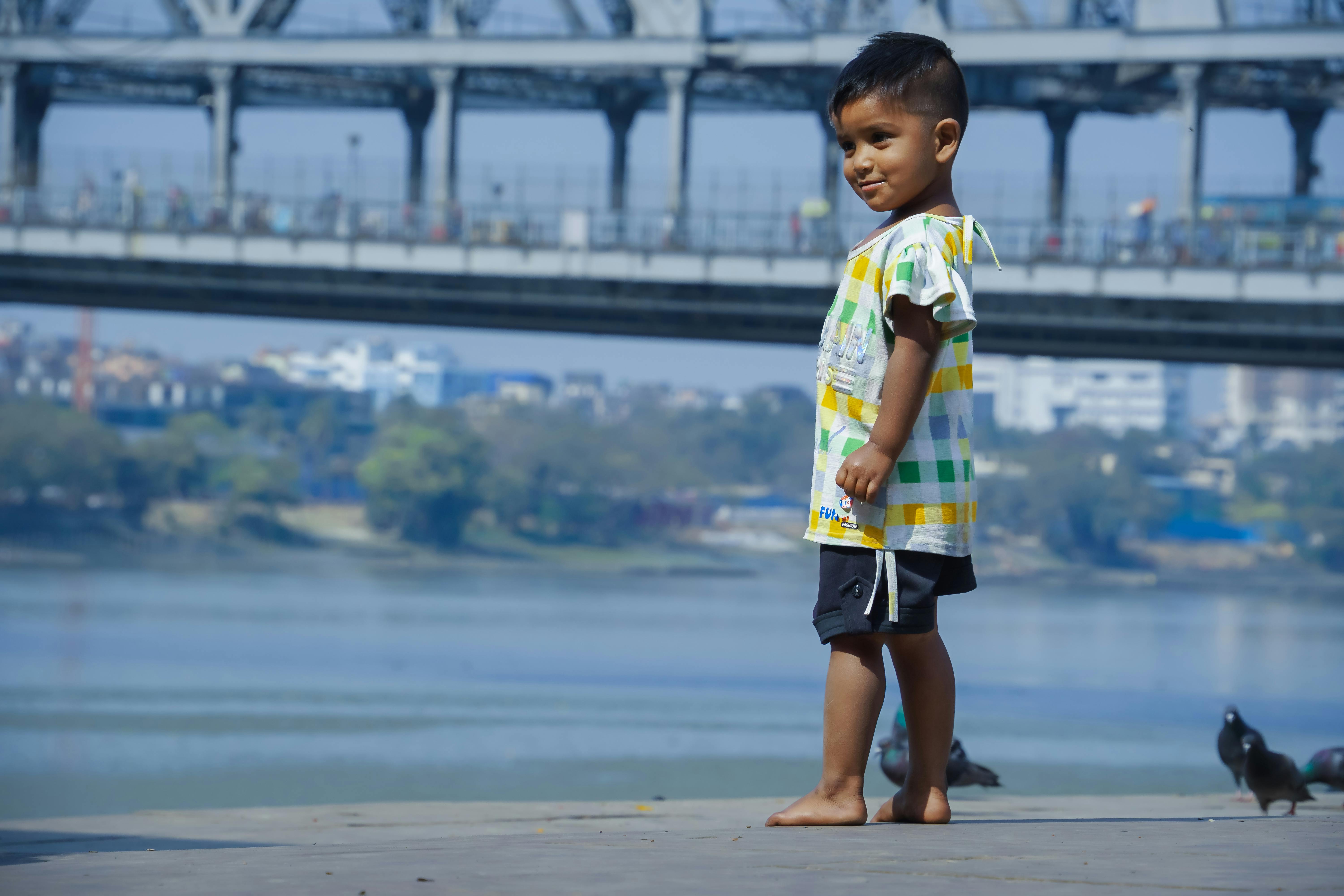 Kostnadsfria En ung pojke står barfota vid floden i Indien med en bro i bakgrunden. Stock foto