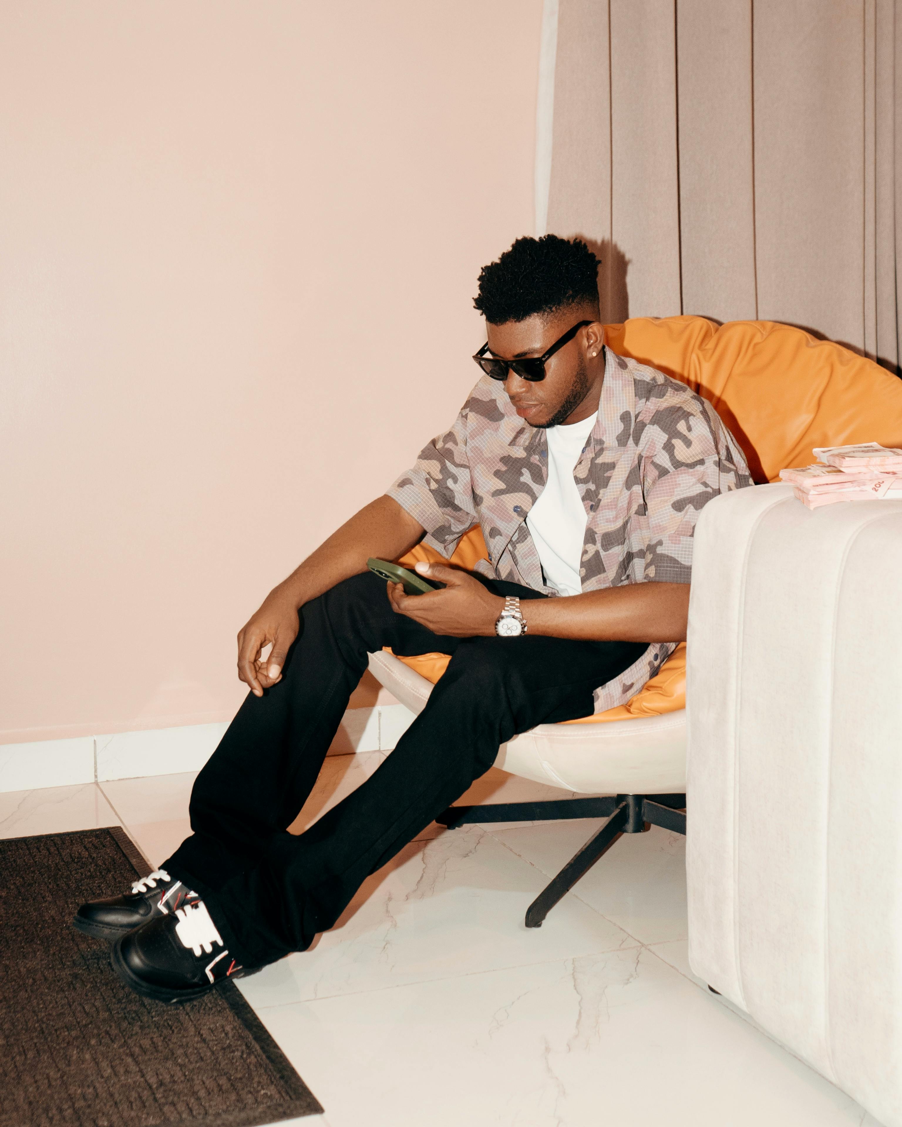 bezplatná Módní muž se slunečními brýlemi sedí uvnitř a v uvolněné atmosféře prohlíží svůj smartphone. Základová fotografie