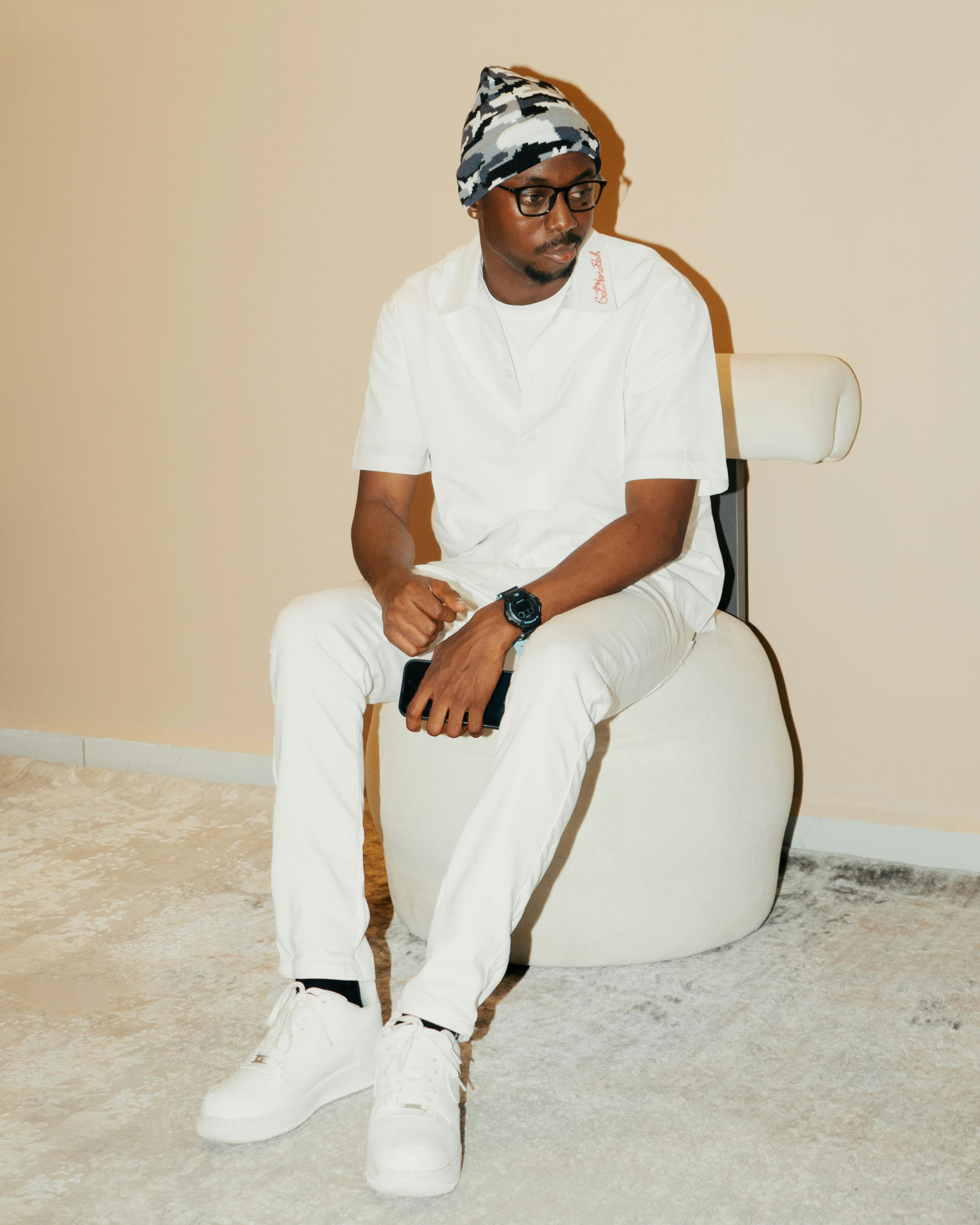 免费 一位身着白色服装、头戴白色帽子的时尚男士坐在室内现代风格的椅子上。 素材图片