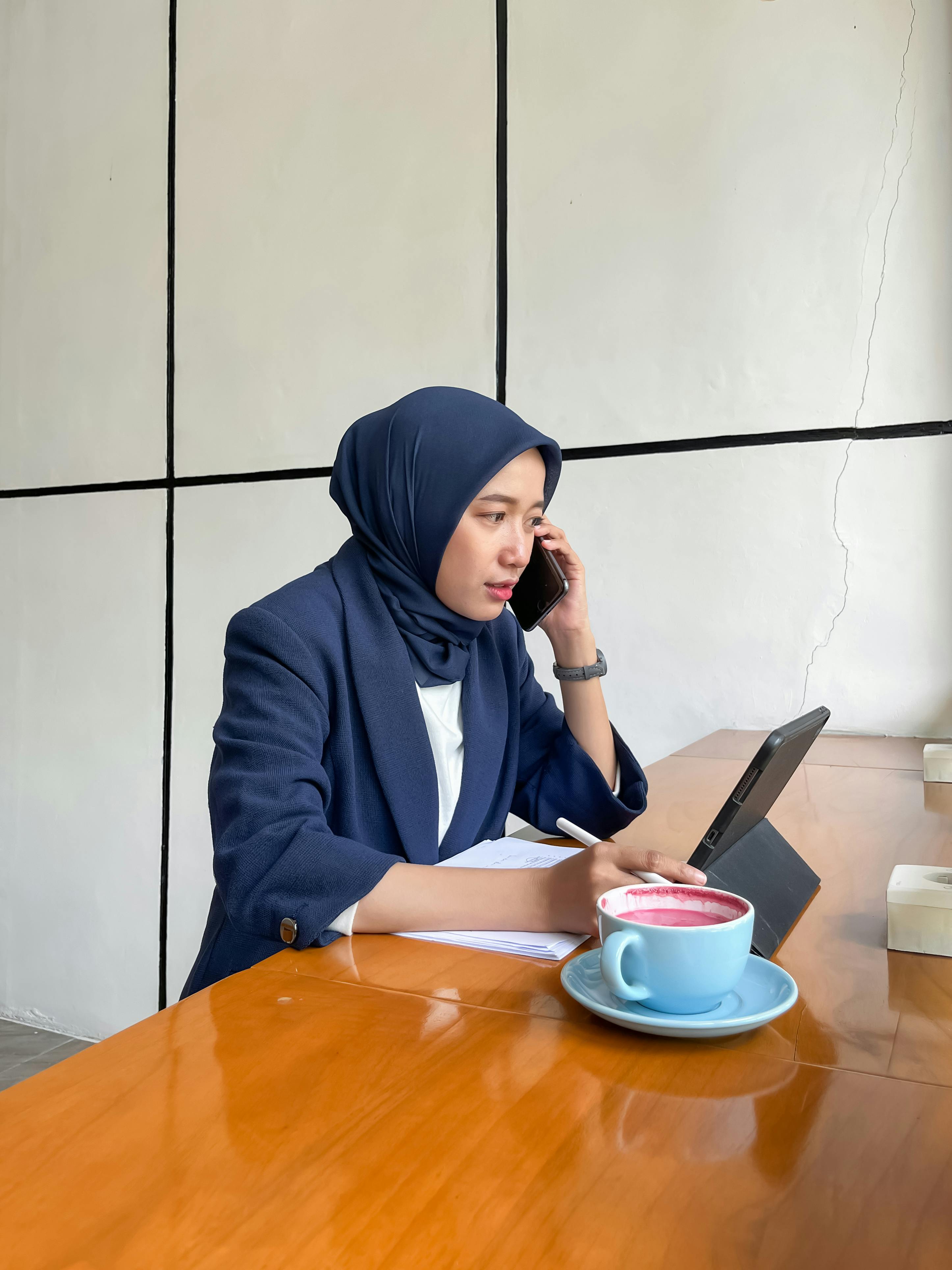 Kostenlos Eine berufstätige Frau mit Hijab erledigt in einem modernen Café-Ambiente mehrere Aufgaben gleichzeitig mit einem Tablet und einer Tasse Kaffee. Stock-Foto