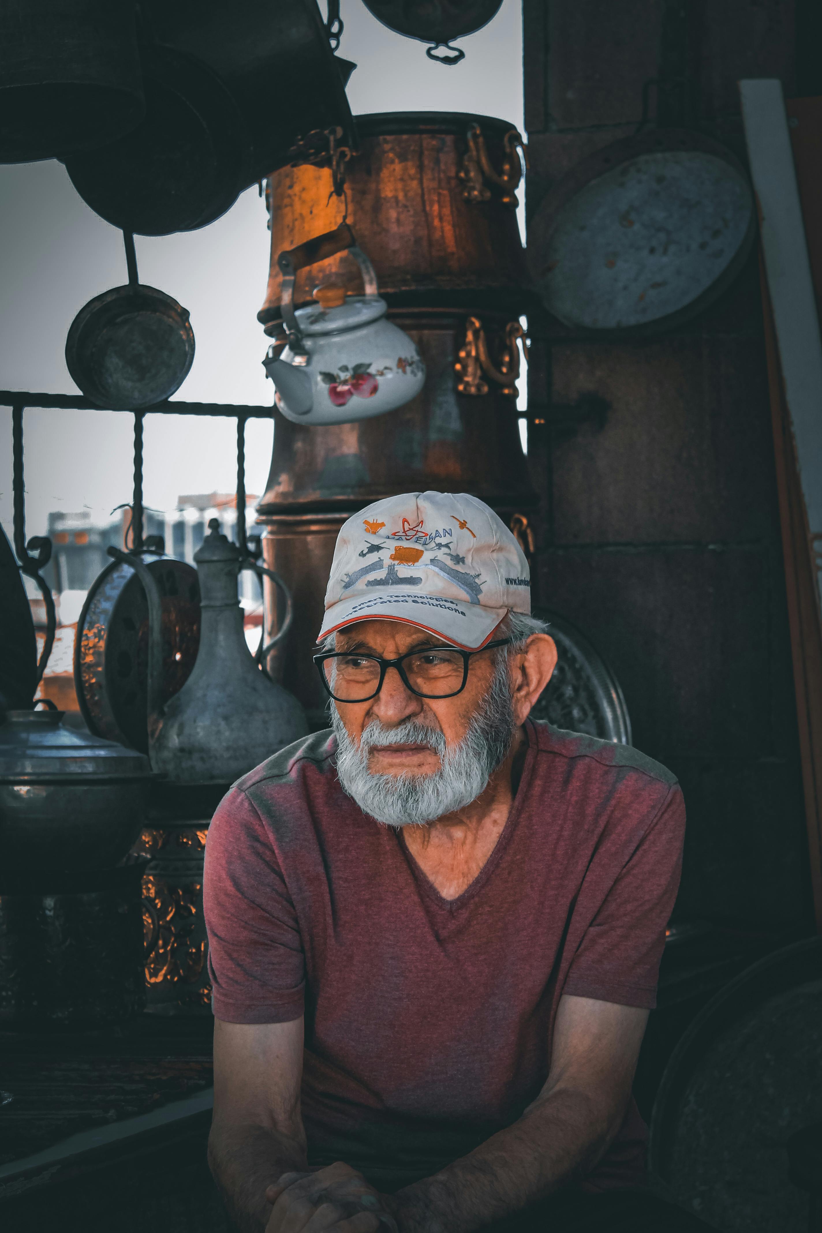 De franc Un home gran amb ulleres i barba seu entre estris de coure vintage en un entorn exterior. Foto d'estoc
