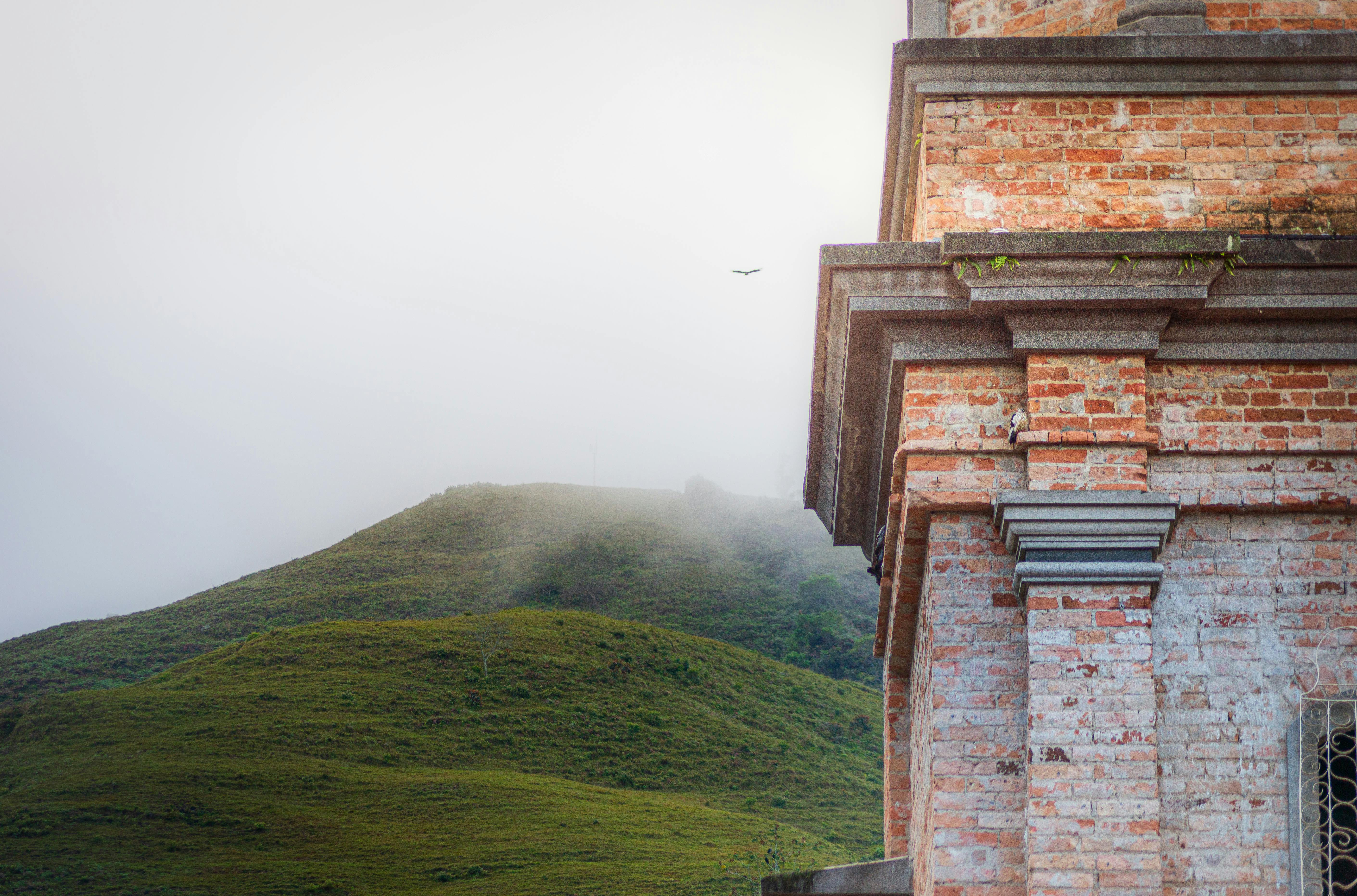 bezplatná Červená cihlová budova stojí před mlhavými zelenými kopci, pod zataženou oblohou. Klidná scenérie. Základová fotografie