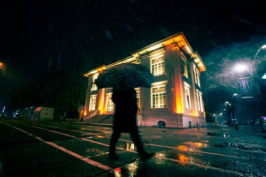 Silhouette of a person with an umbrella passing by an illuminated building on a rainy night.