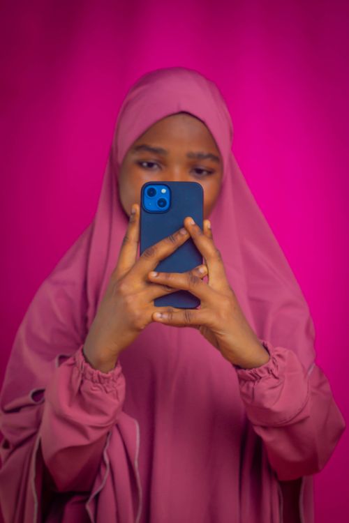 Free A young woman wearing a pink hijab takes a selfie using a smartphone against a vibrant pink backdrop. Stock Photo