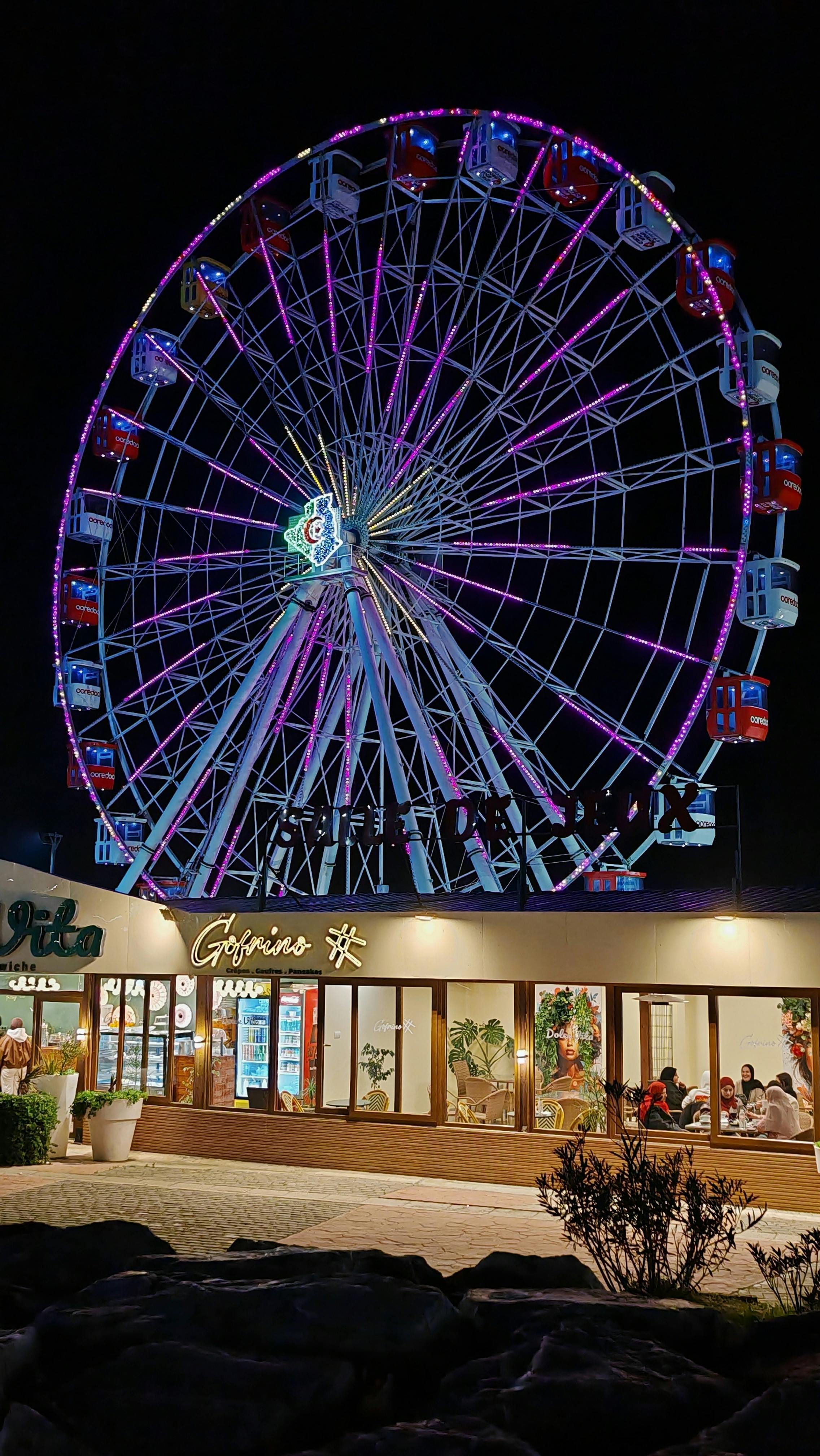 Gratis Una colorata ruota panoramica illuminata stagliandosi contro il cielo notturno, sopra una vivace zona ricca di ristoranti. Foto a disposizione