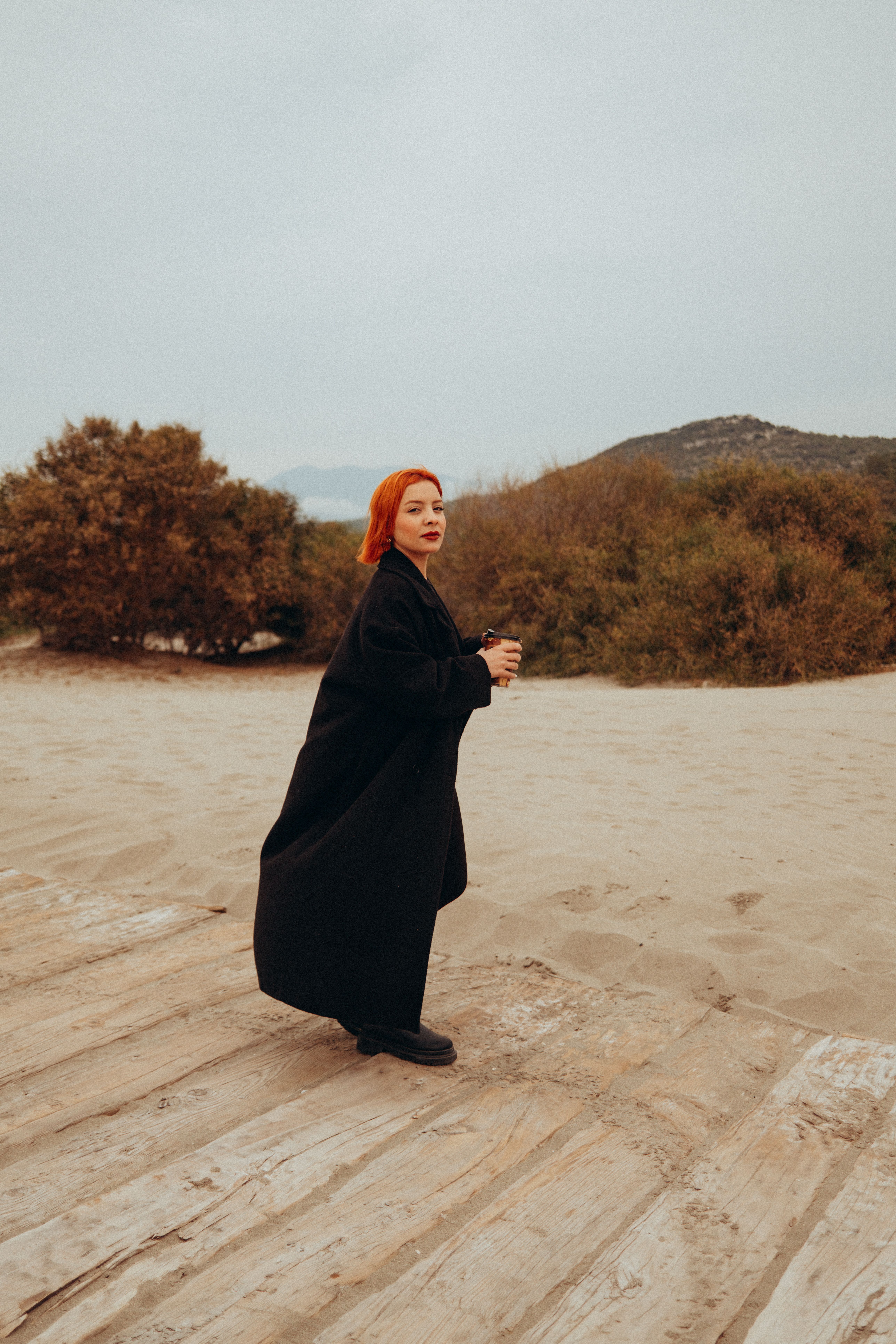 A woman with red hair in a black coat walks on a sandy path, surrounded by nature. Outdoor casual scene.