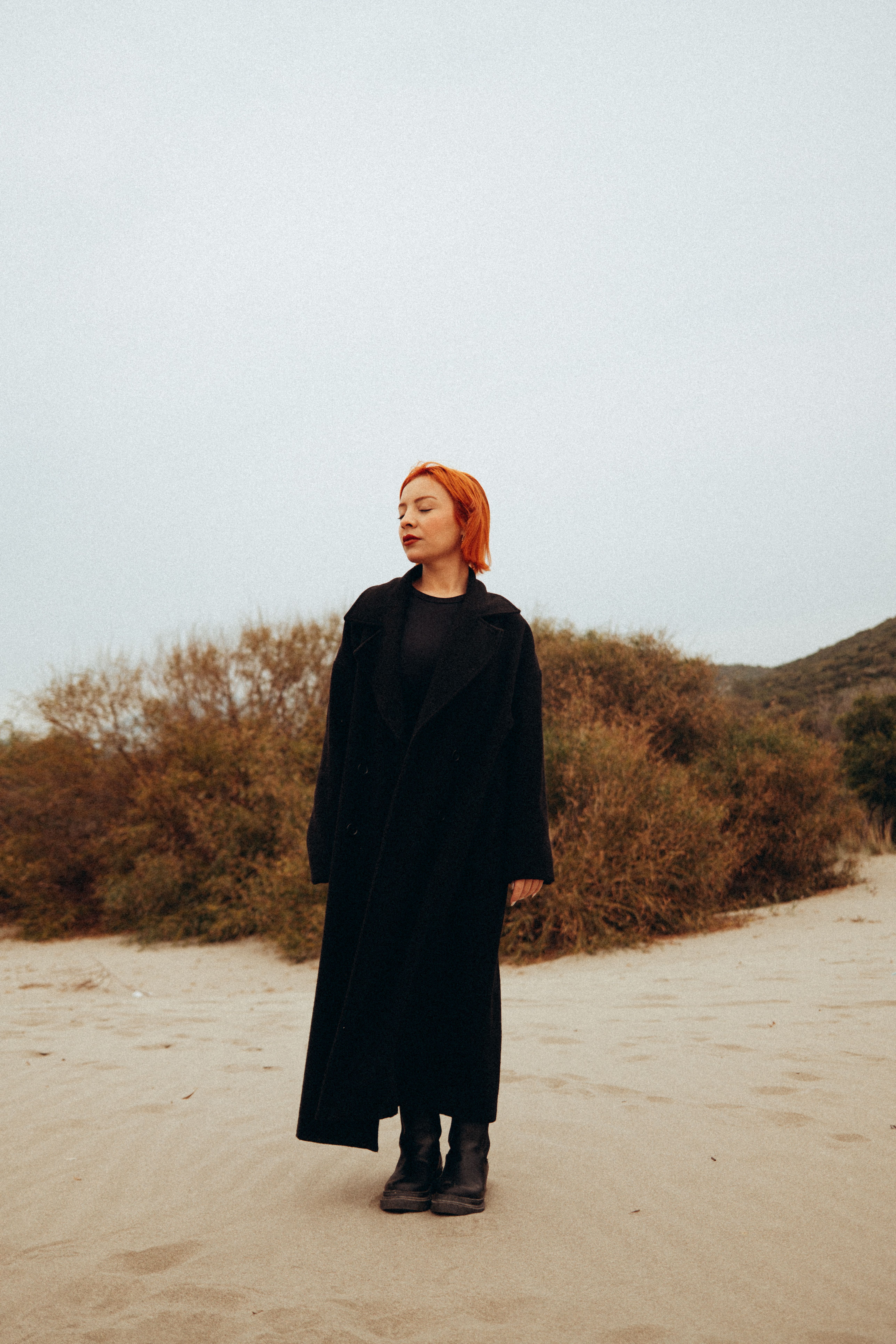 Woman with red hair in a black coat stands on a serene sandy beach.