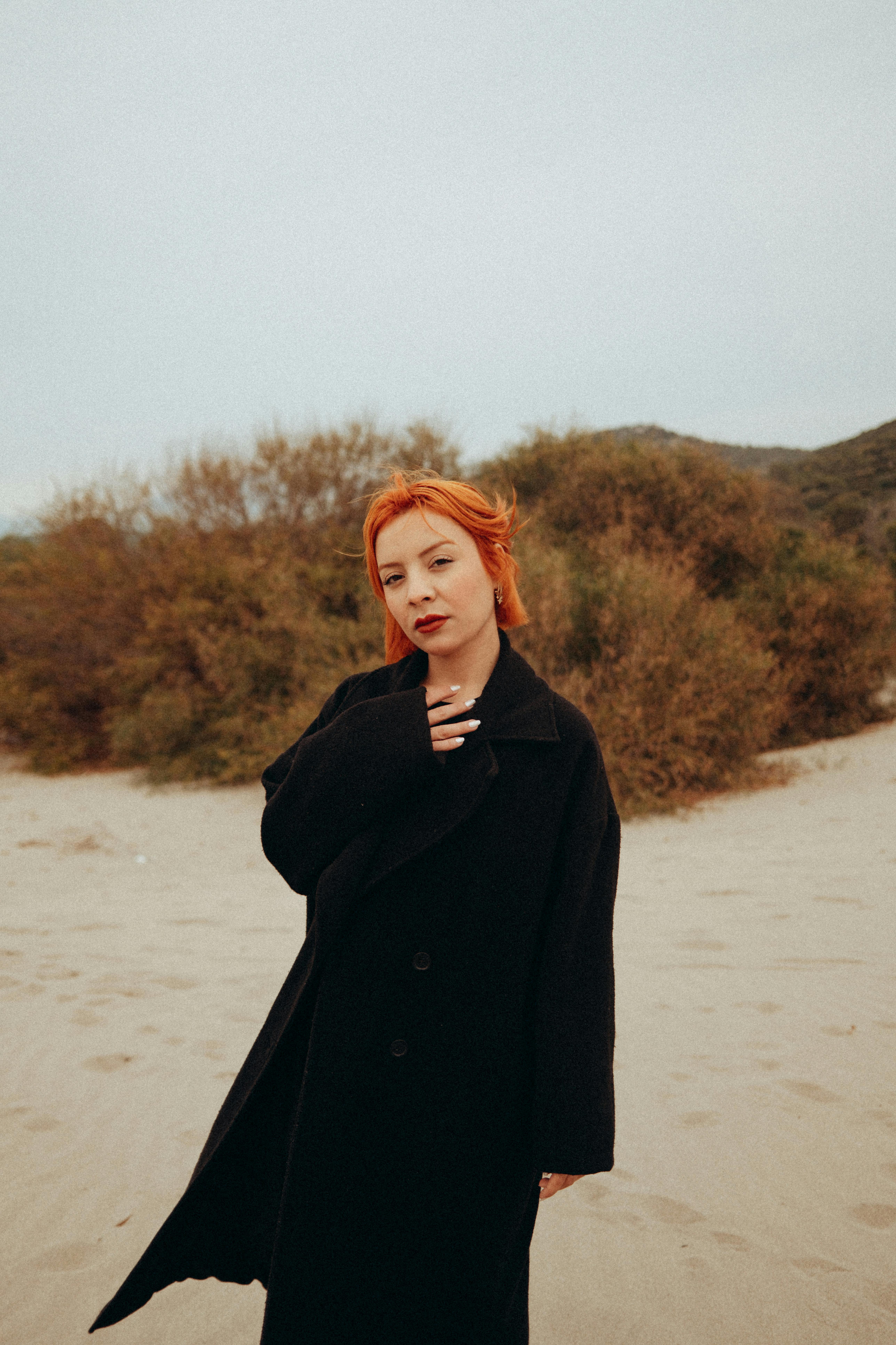 grátis Retrato de uma mulher ruiva em pé numa praia de areia, vestindo um casaco preto, exalando confiança e estilo. Foto profissional