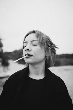 Dramatic black and white portrait of a woman smoking with eyes closed in an outdoor setting.