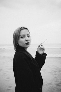 A woman with a cigarette at the beach in a moody black and white photo.