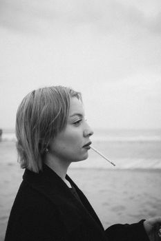A black and white portrait of a woman with short hair smoking by the beach.