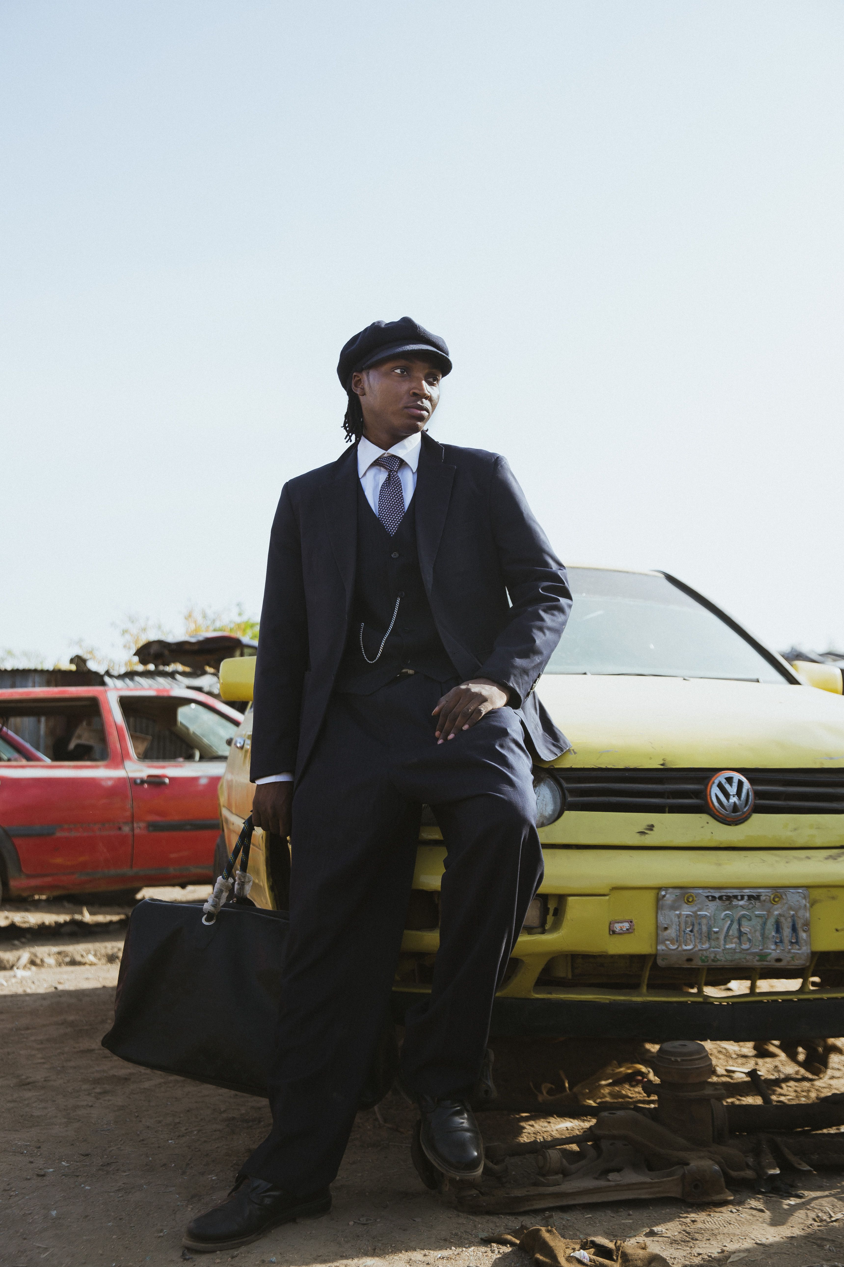 A fashionable man in a classic suit stands confidently against a vintage yellow car in a junkyard setting.