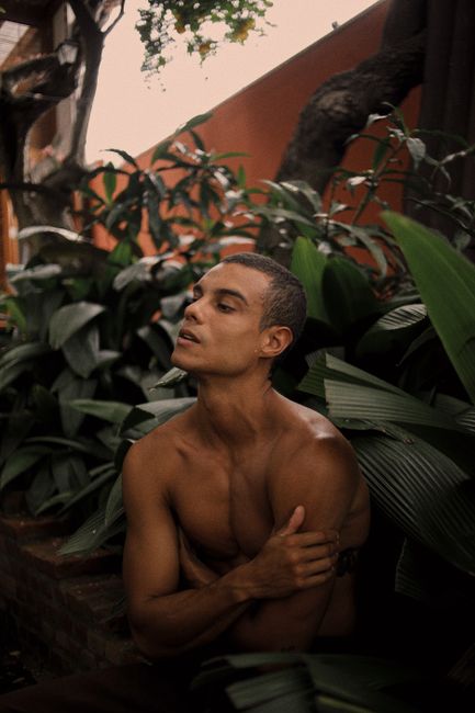 A shirtless man poses among lush tropical plants outdoors in São Paulo.
