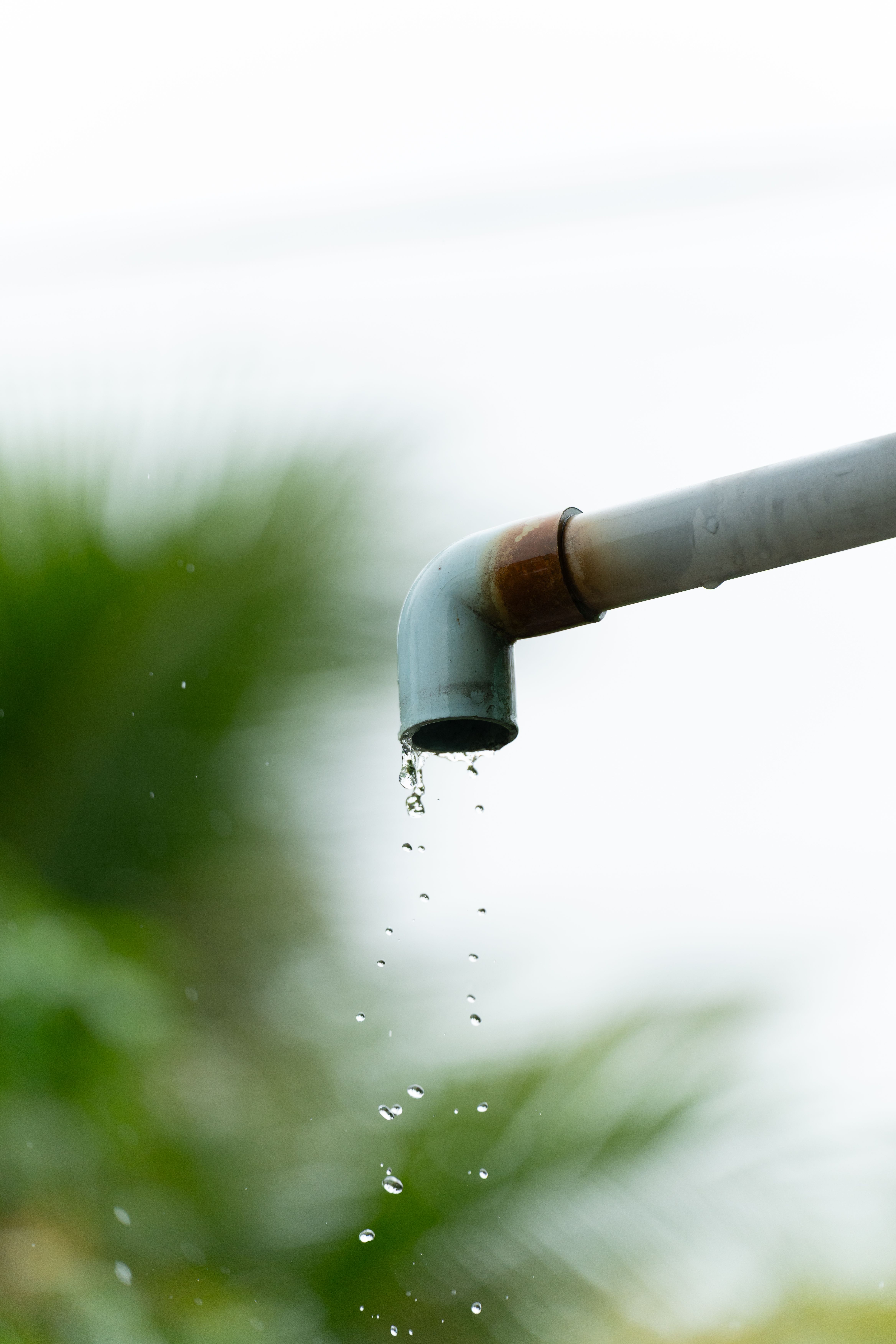 A close-up of water droplets falling from a rusty outdoor pipe against a blurred natural background.