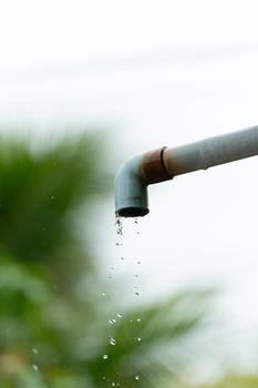 A close-up of water droplets falling from a rusty outdoor pipe against a blurred natural background.