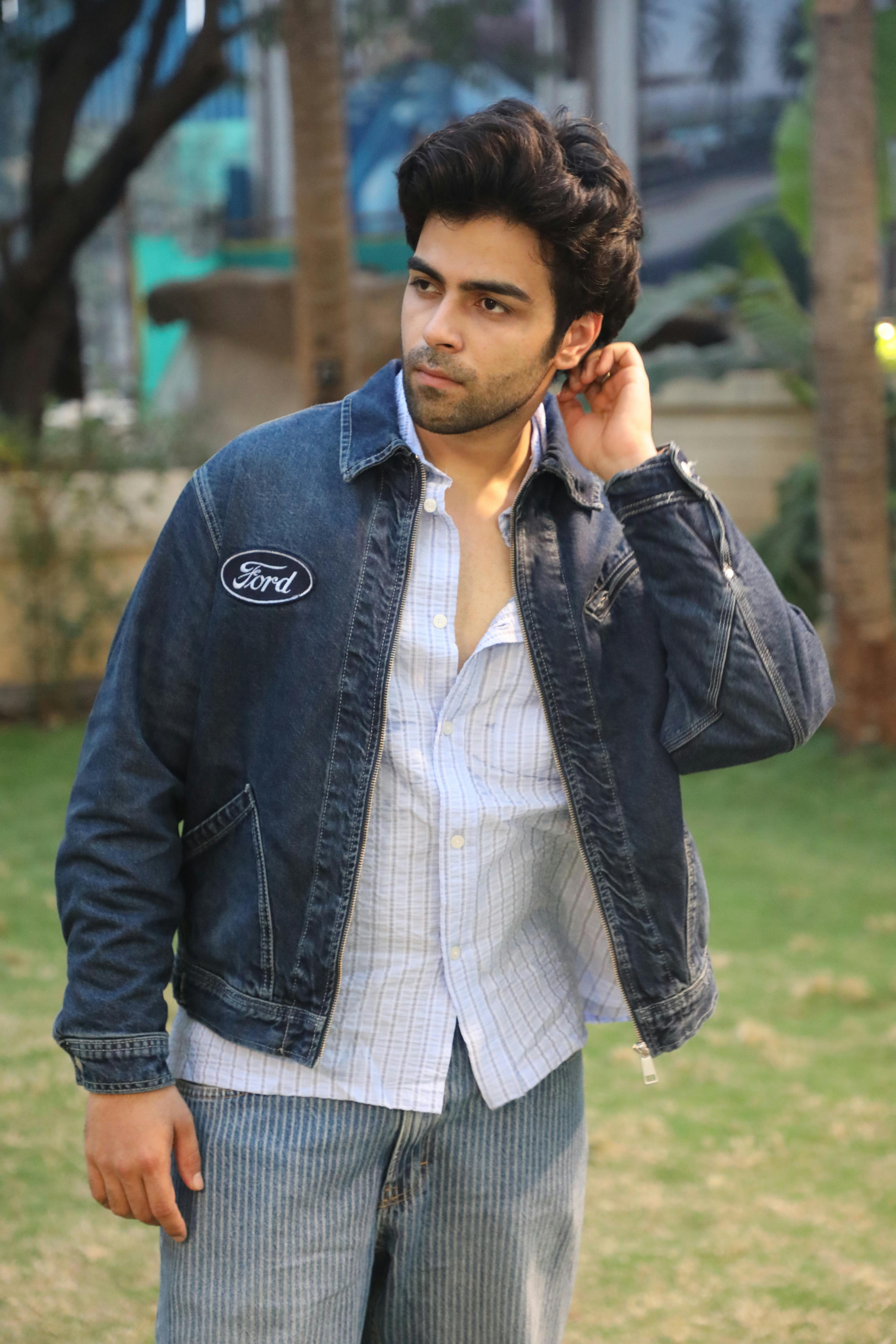 A young man in a denim jacket posing naturally in an outdoor setting.