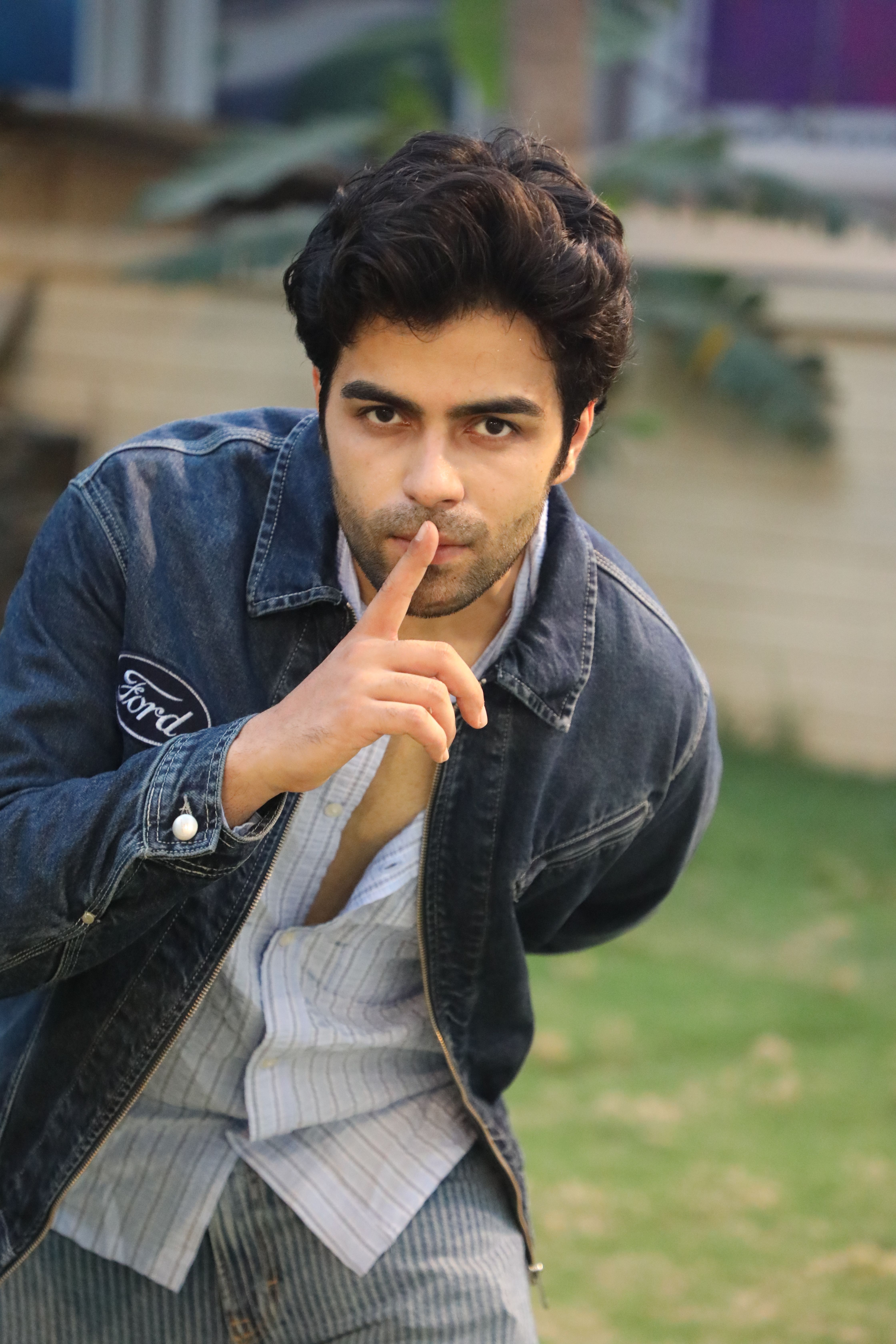 A young man outdoors in a denim jacket gesturing for silence, surrounded by greenery.