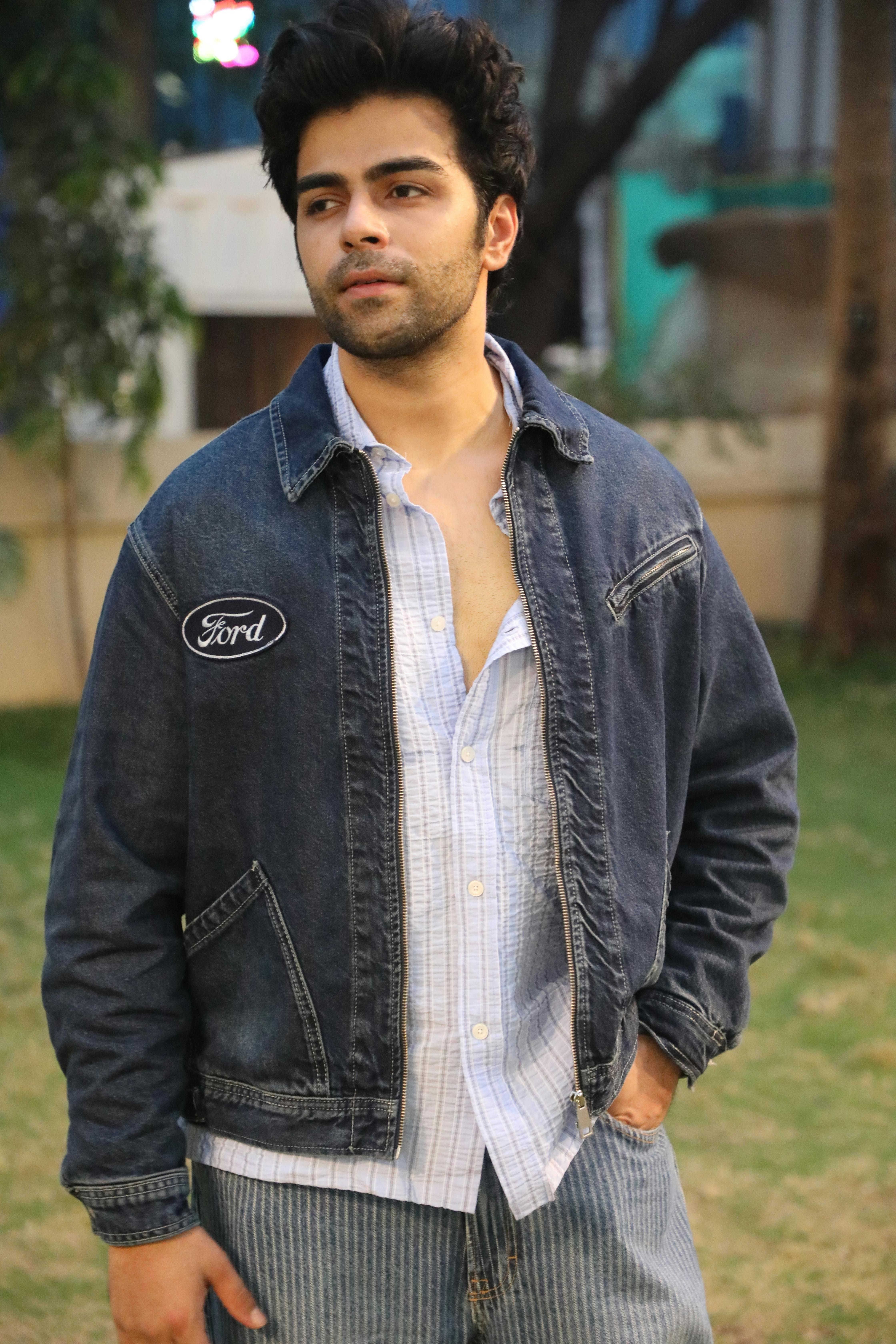Young man wearing denim jacket and jeans looking thoughtful in a garden setting.