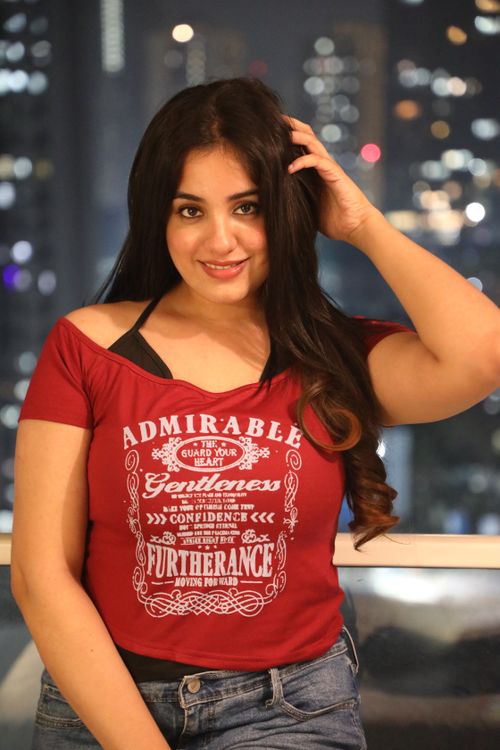 Free A young woman in a red shirt smiles with city lights in the background at night. Stock Photo